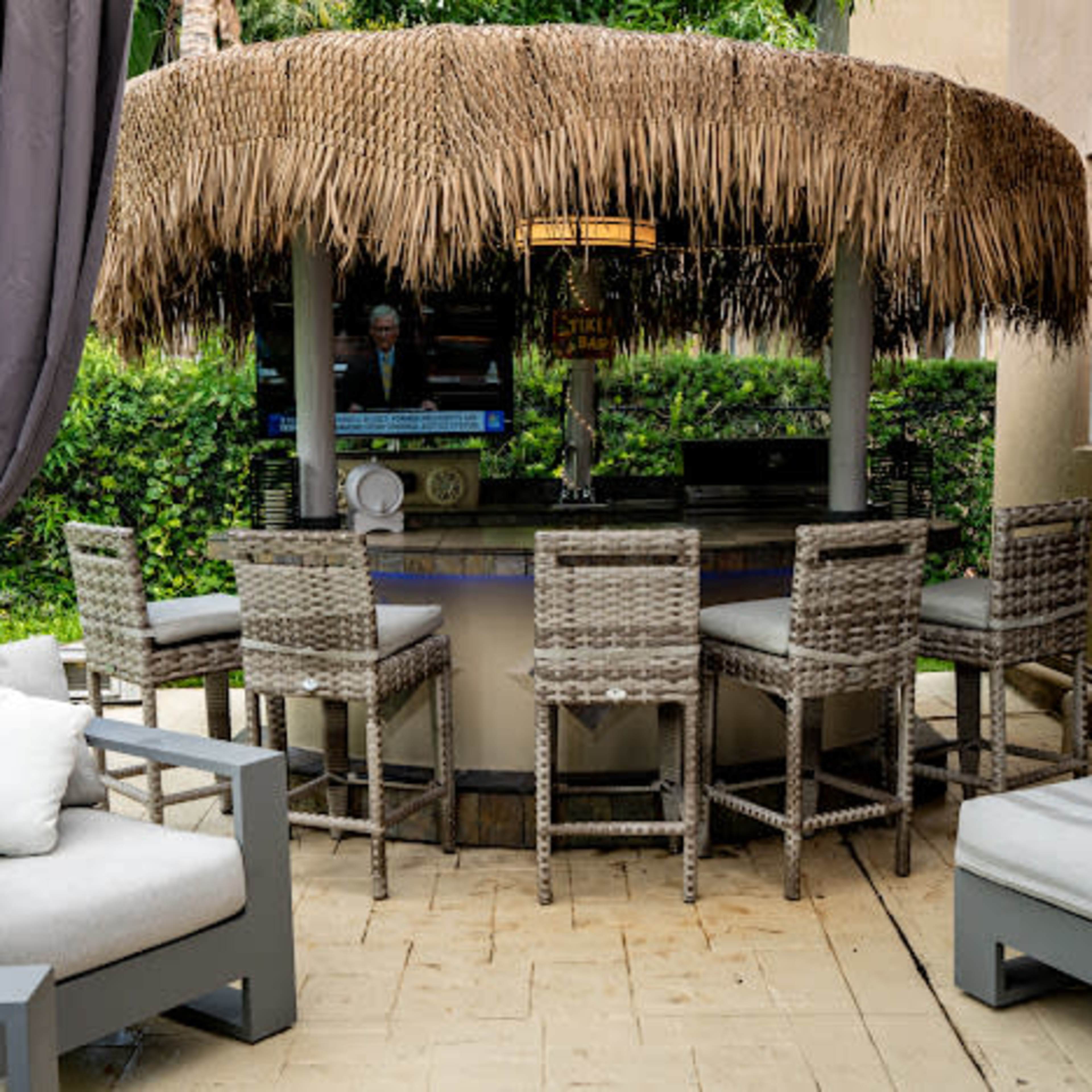 An outdoor bar with a thatched roof surrounded by gray wicker bar stools and a television in the background.