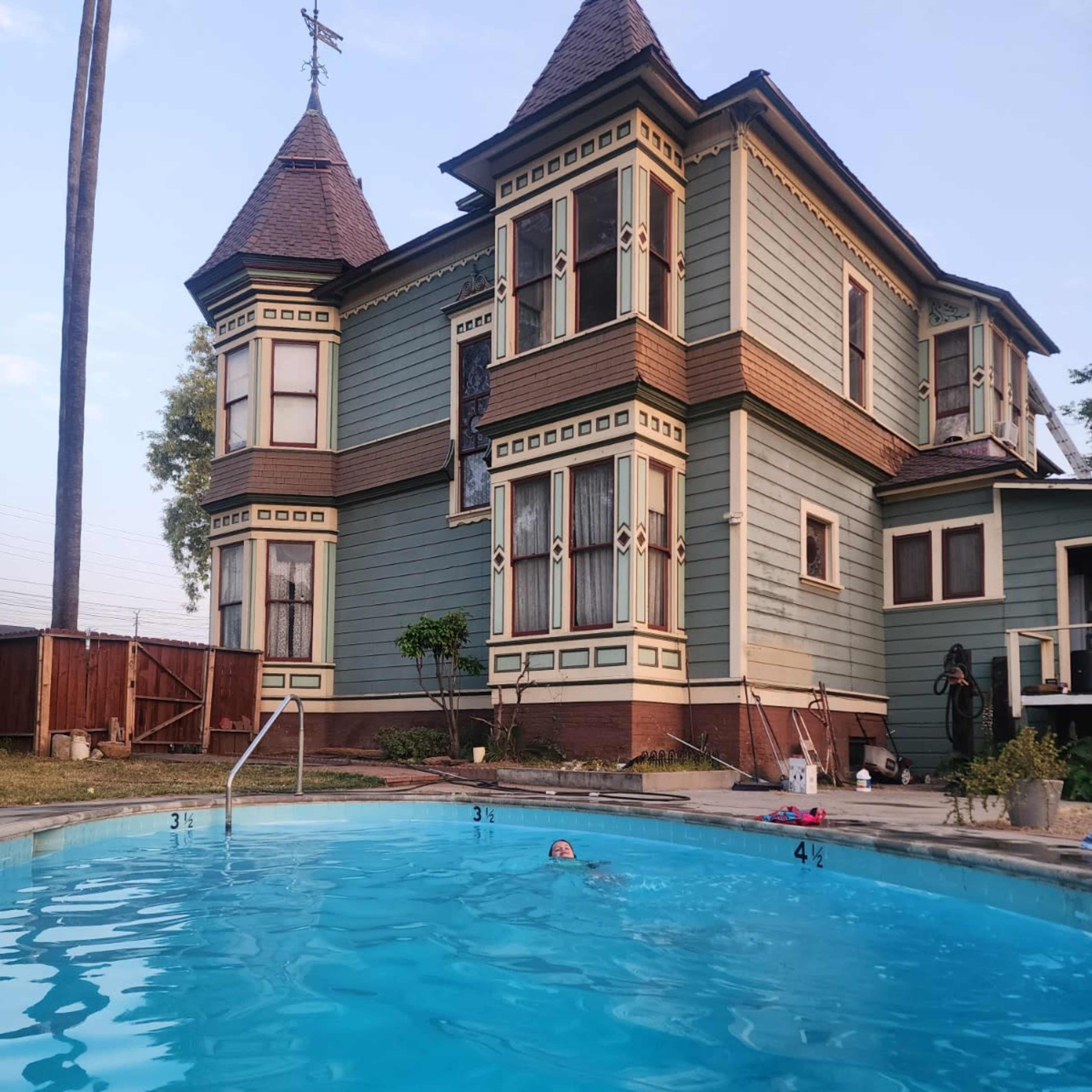 A person swims in a clear blue pool in front of a large, historic two-story house with Victorian architectural features.