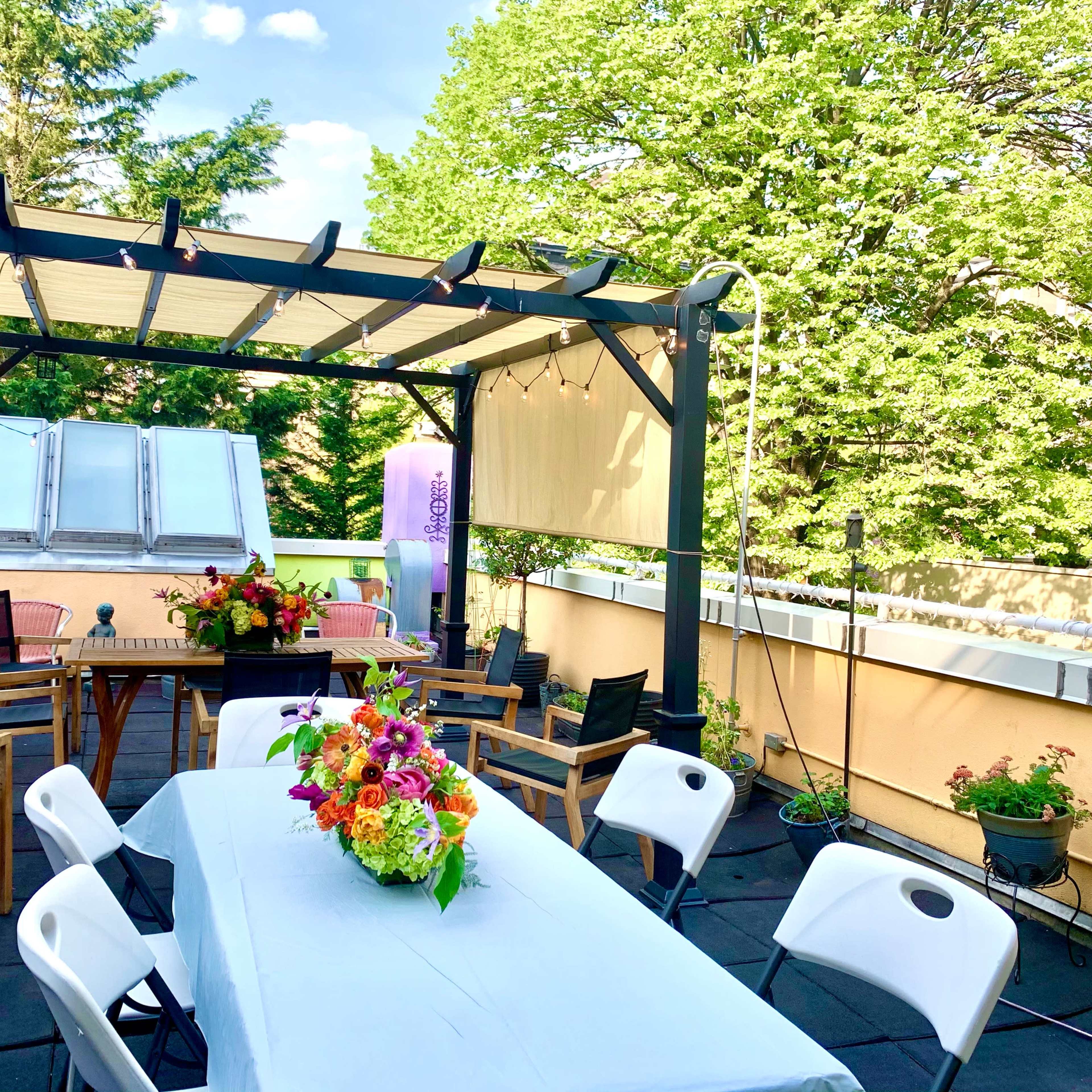 The rooftop garden features a pergola with seating, a large table adorned with flowers, and various plants in pots along the railing.