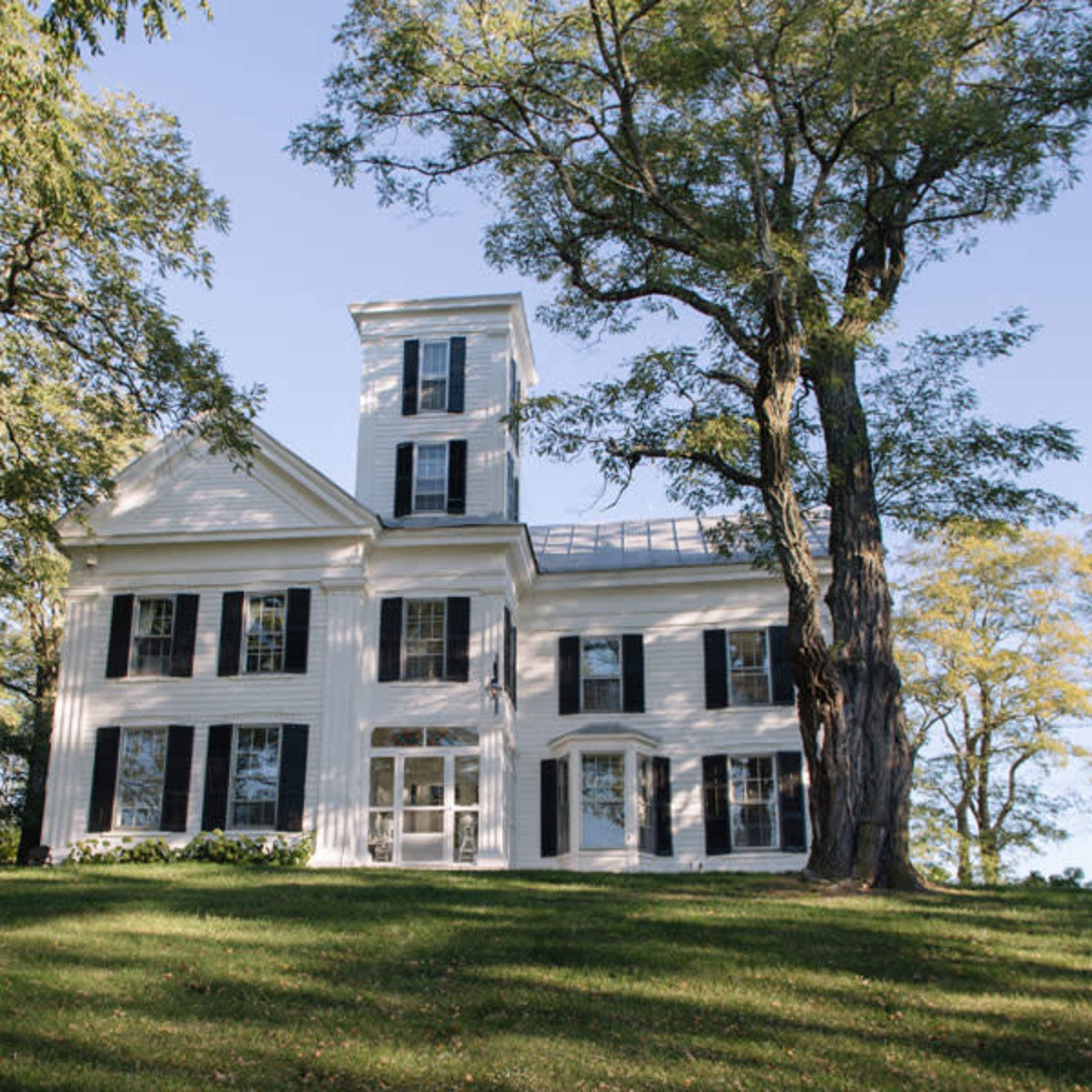 A large white house with black shutters stands on a grassy hill, surrounded by tall trees.