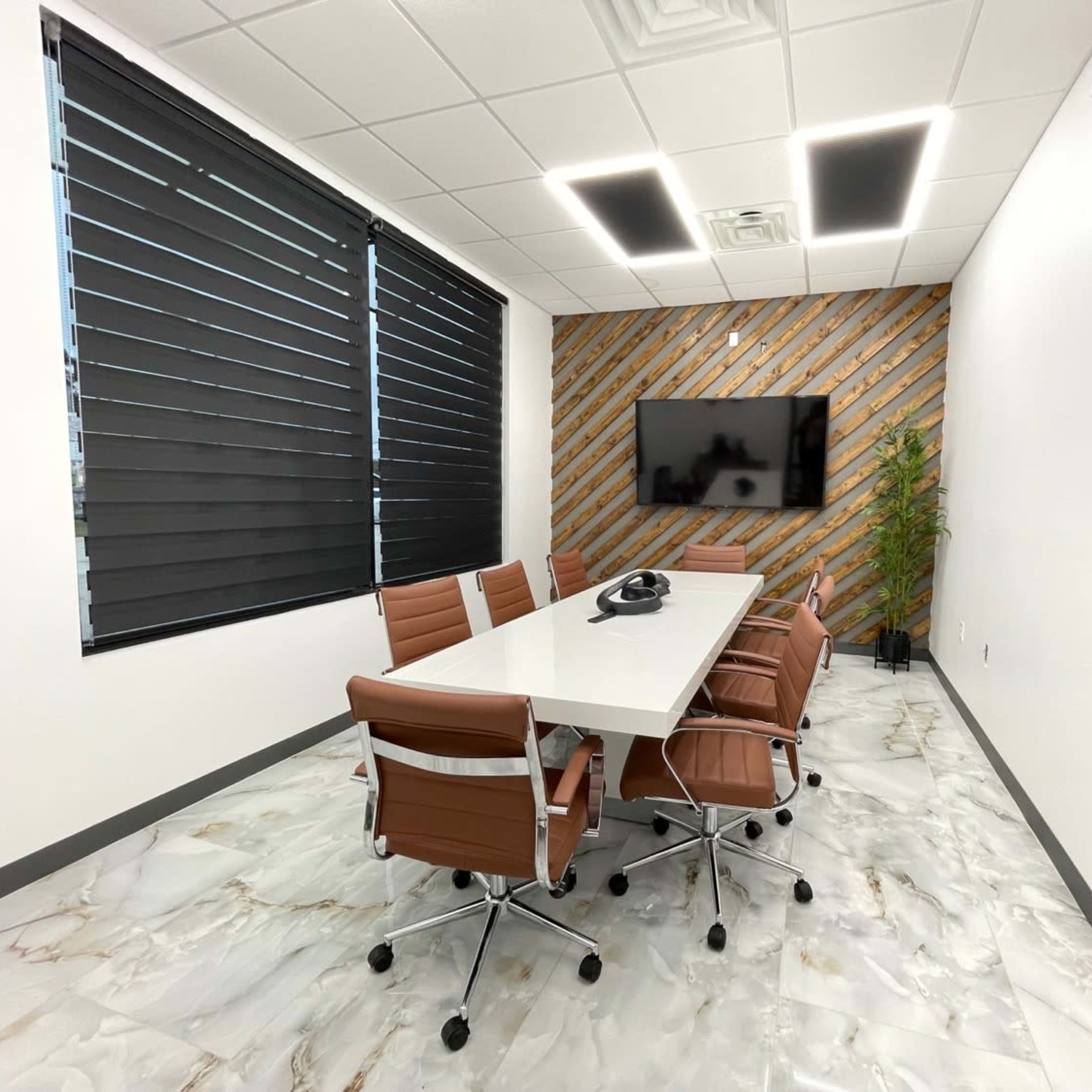 A modern conference room with a long white table, ergonomic chairs, a wall-mounted TV, and wooden accent wall panels.
