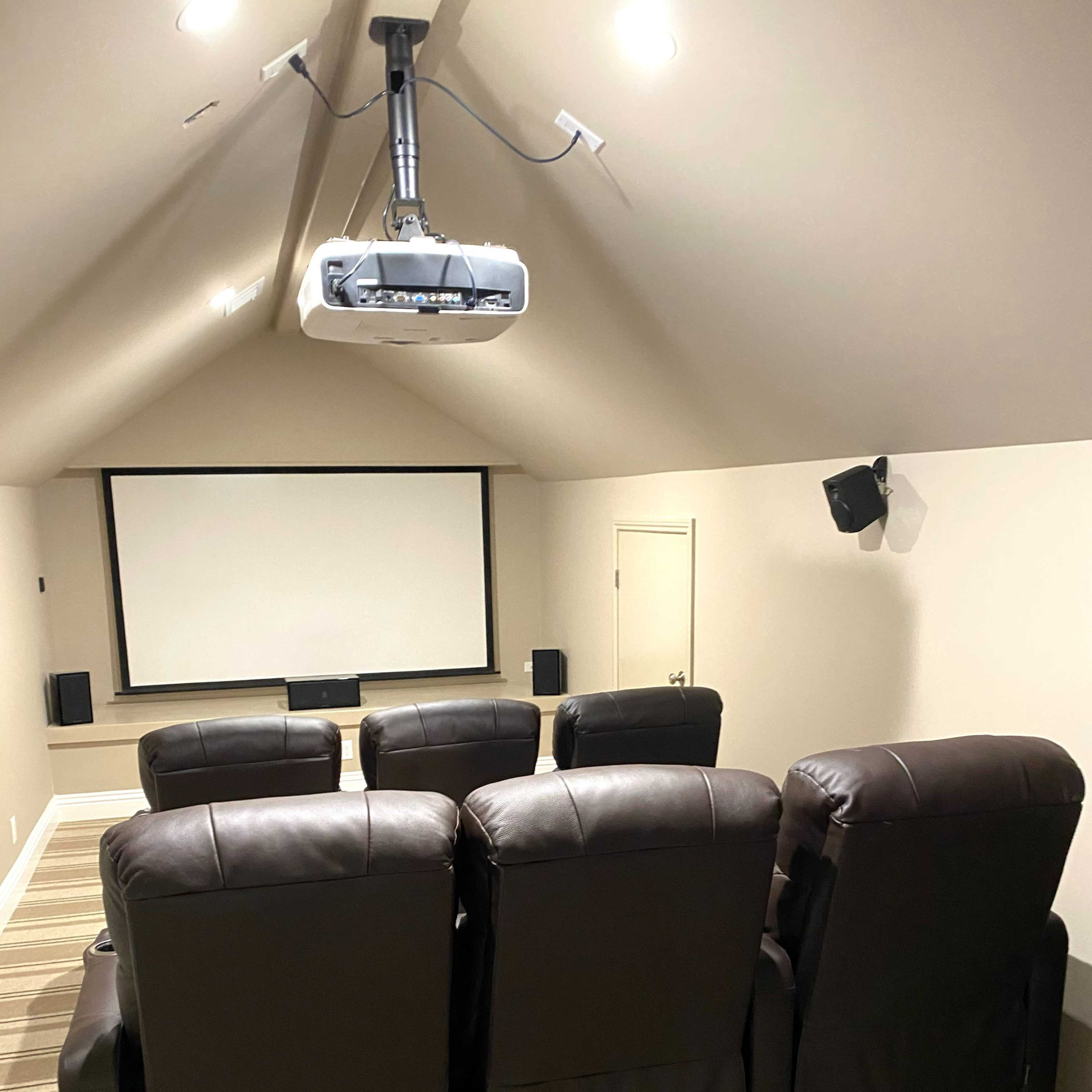 A home theater room with dark leather recliners facing a large projection screen and a ceiling-mounted projector.