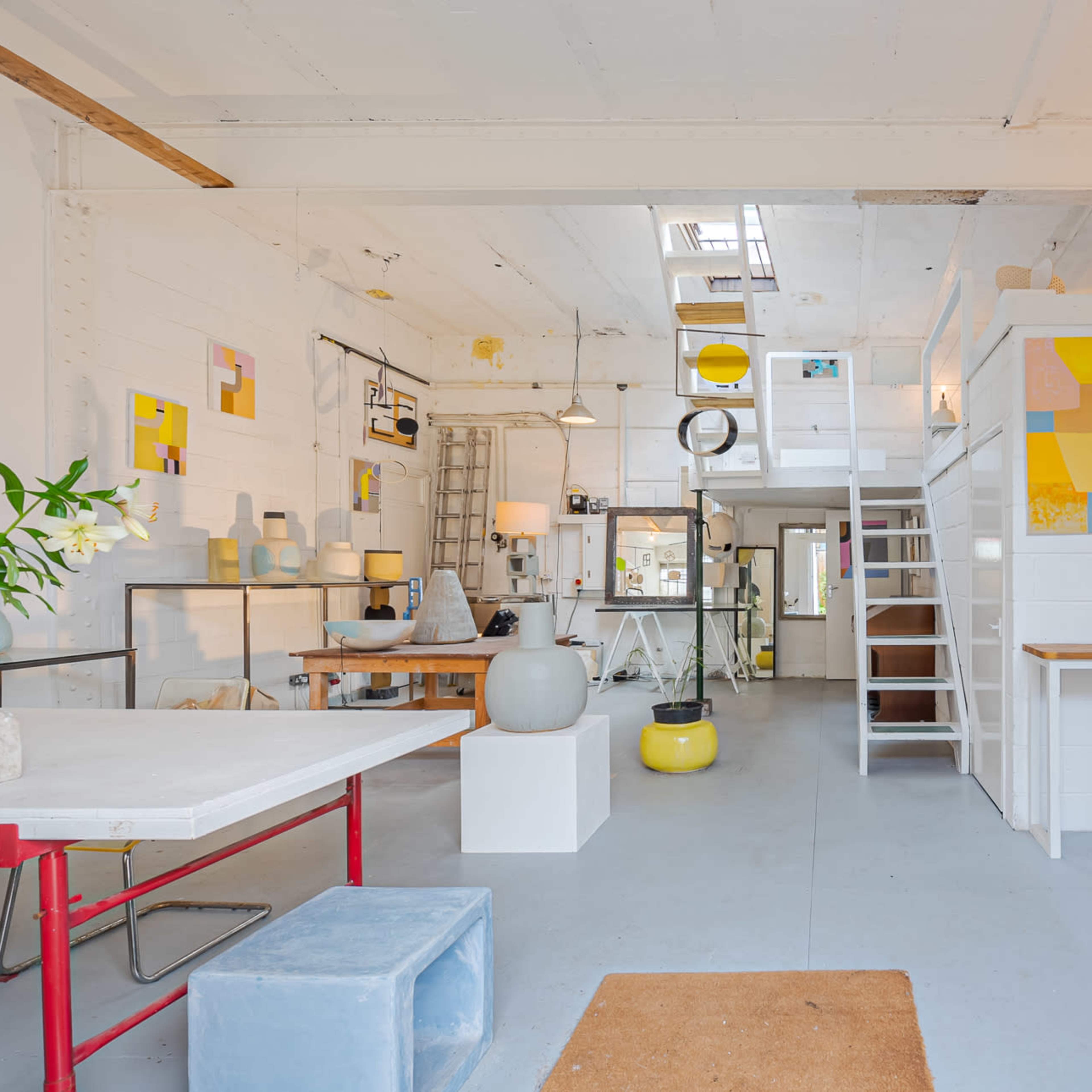 A spacious, well-lit studio with various artworks displayed on the walls and a mix of furniture, including tables, stools, and decorative items.