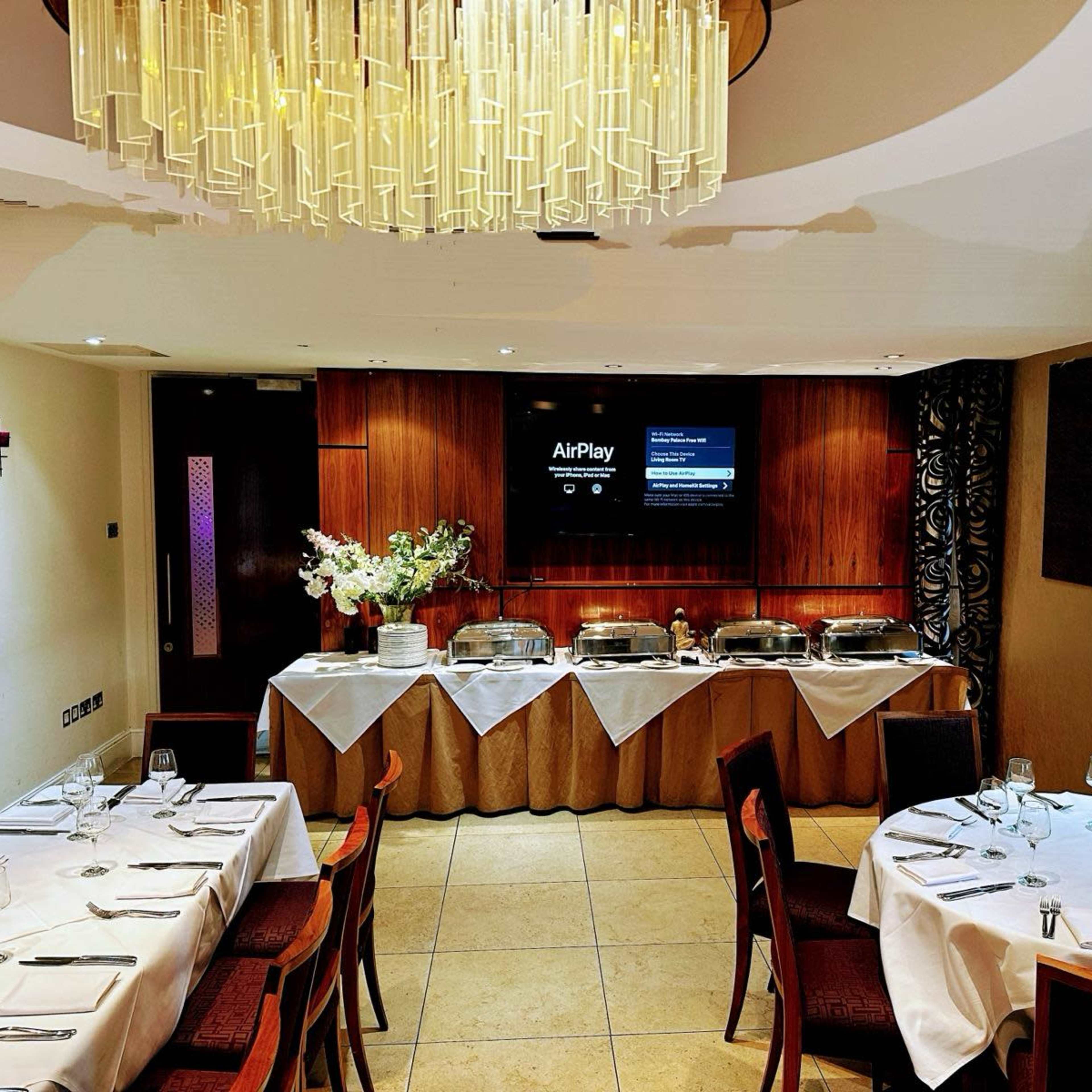A dining room set for an event with tables neatly covered in white tablecloths and a buffet setup featuring chafing dishes against a backdrop of a television screen and decorative lighting.