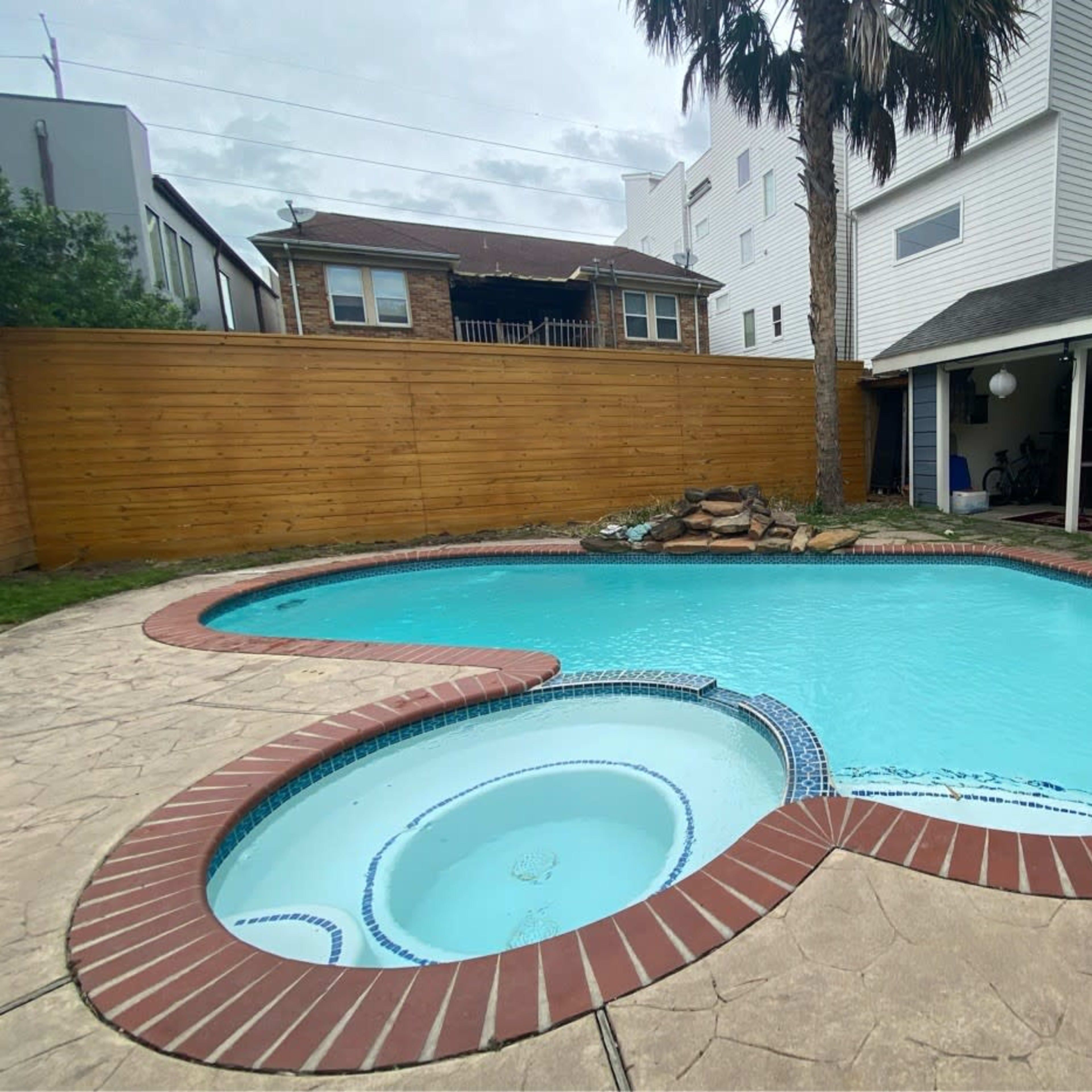A turquoise-shaped pool surrounded by a stone patio and a wooden fence, with a small outdoor structure in the background.