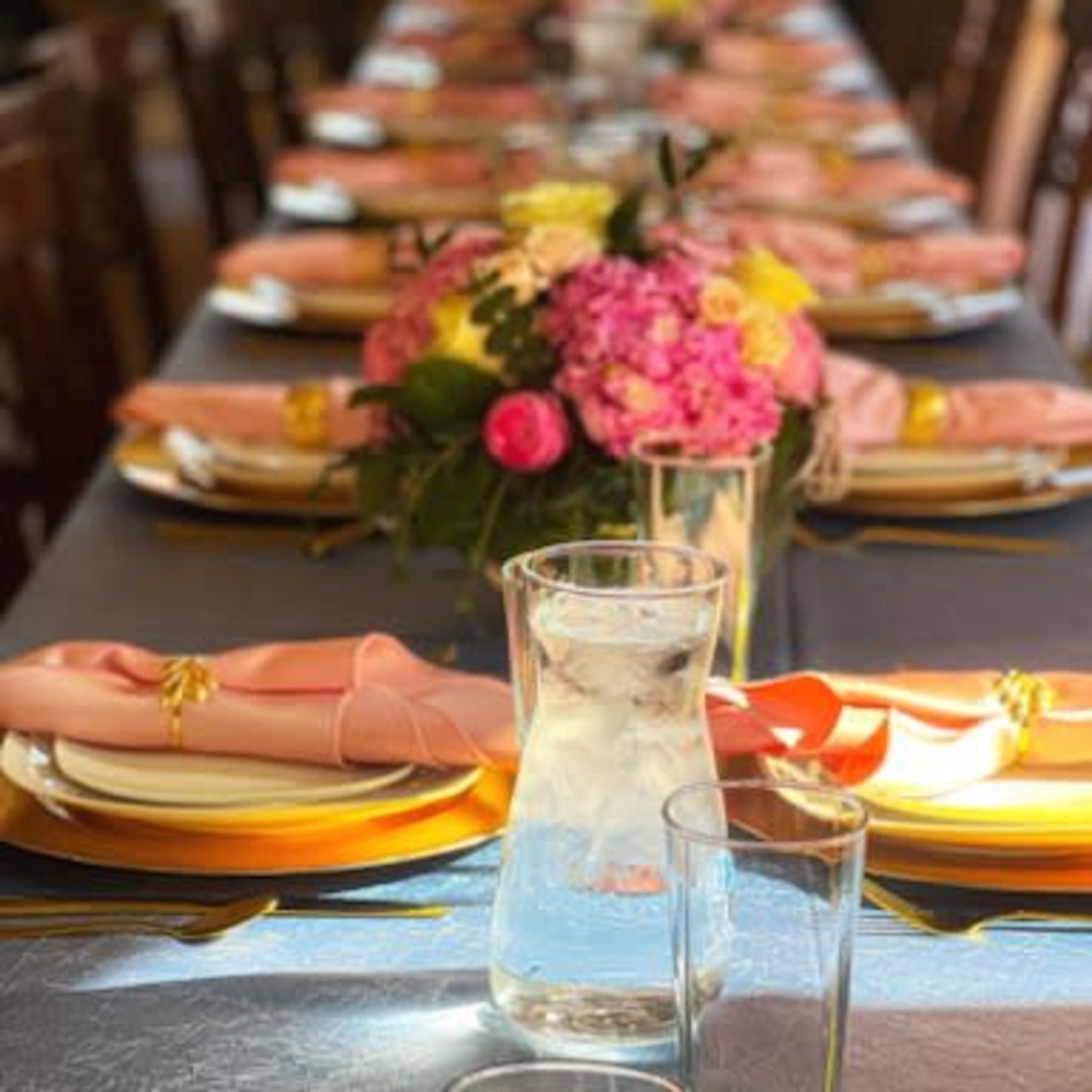 A long dining table is set with neatly arranged plates, glasses, and floral centerpieces surrounded by wooden chairs.