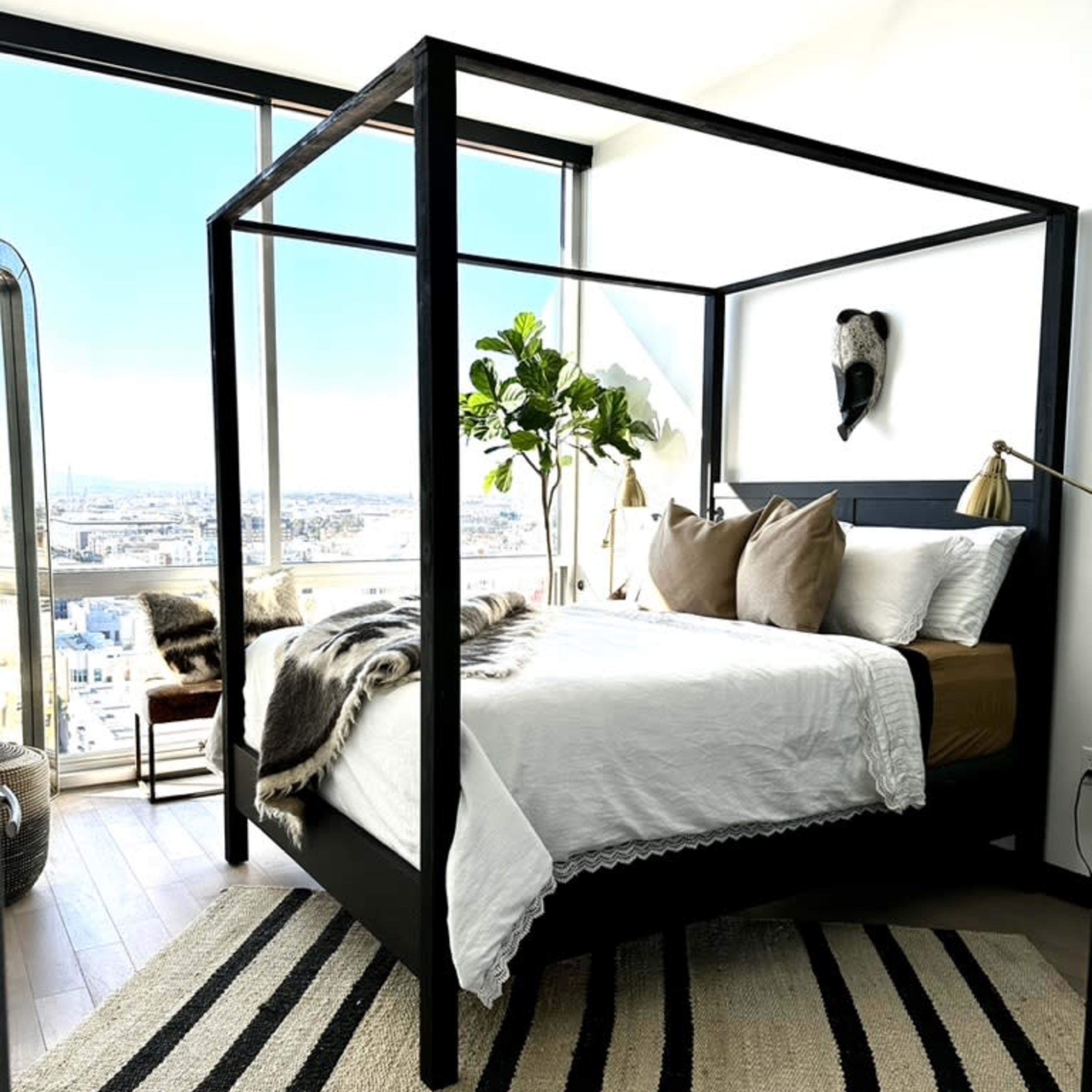 A modern bedroom features a black four-poster bed with a white bedding set, a large window offering a city view, and a striped rug on the wooden floor.