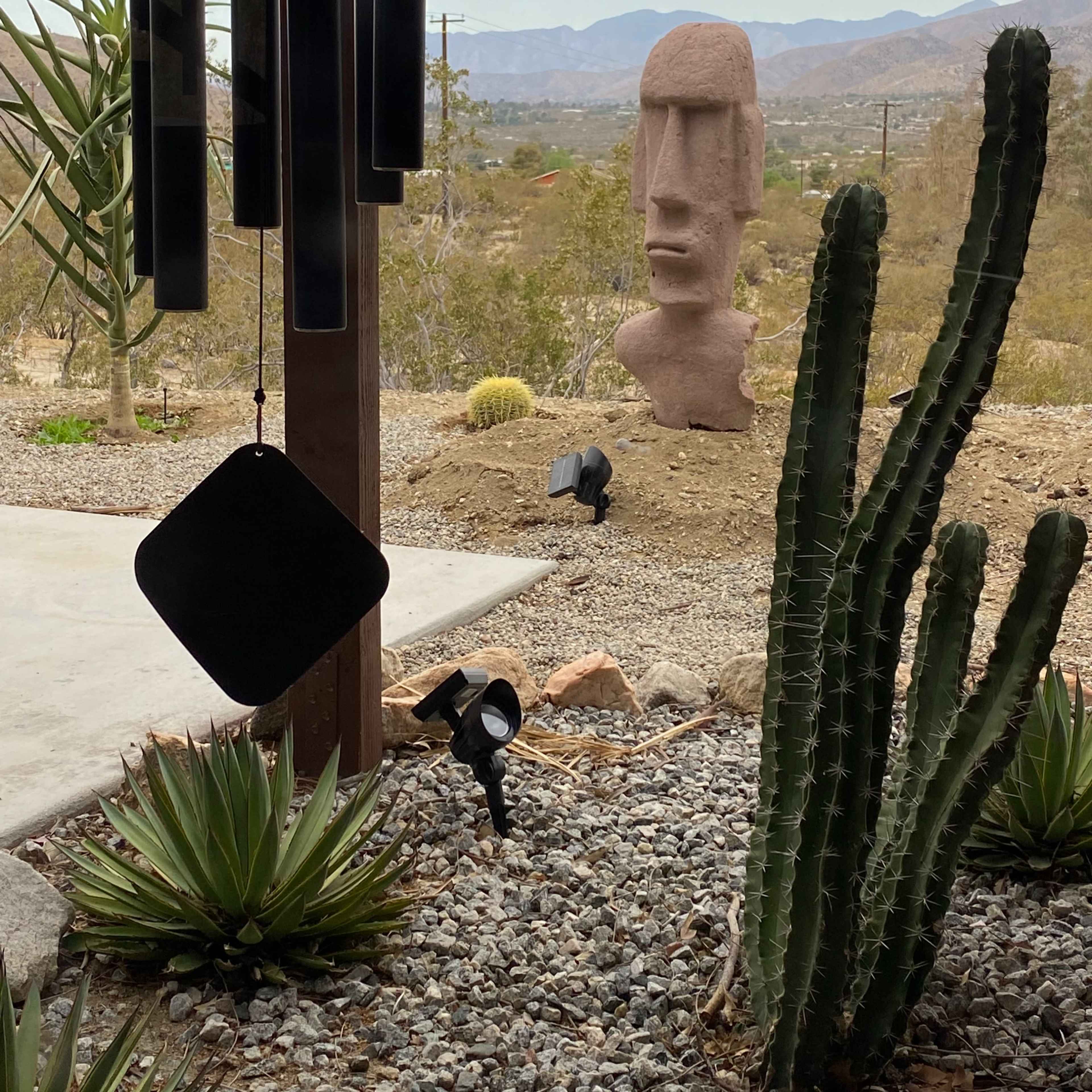 A wind chime in the foreground with a large stone statue, resembling an Easter Island head, positioned in a desert landscape surrounded by cacti and distant mountains.