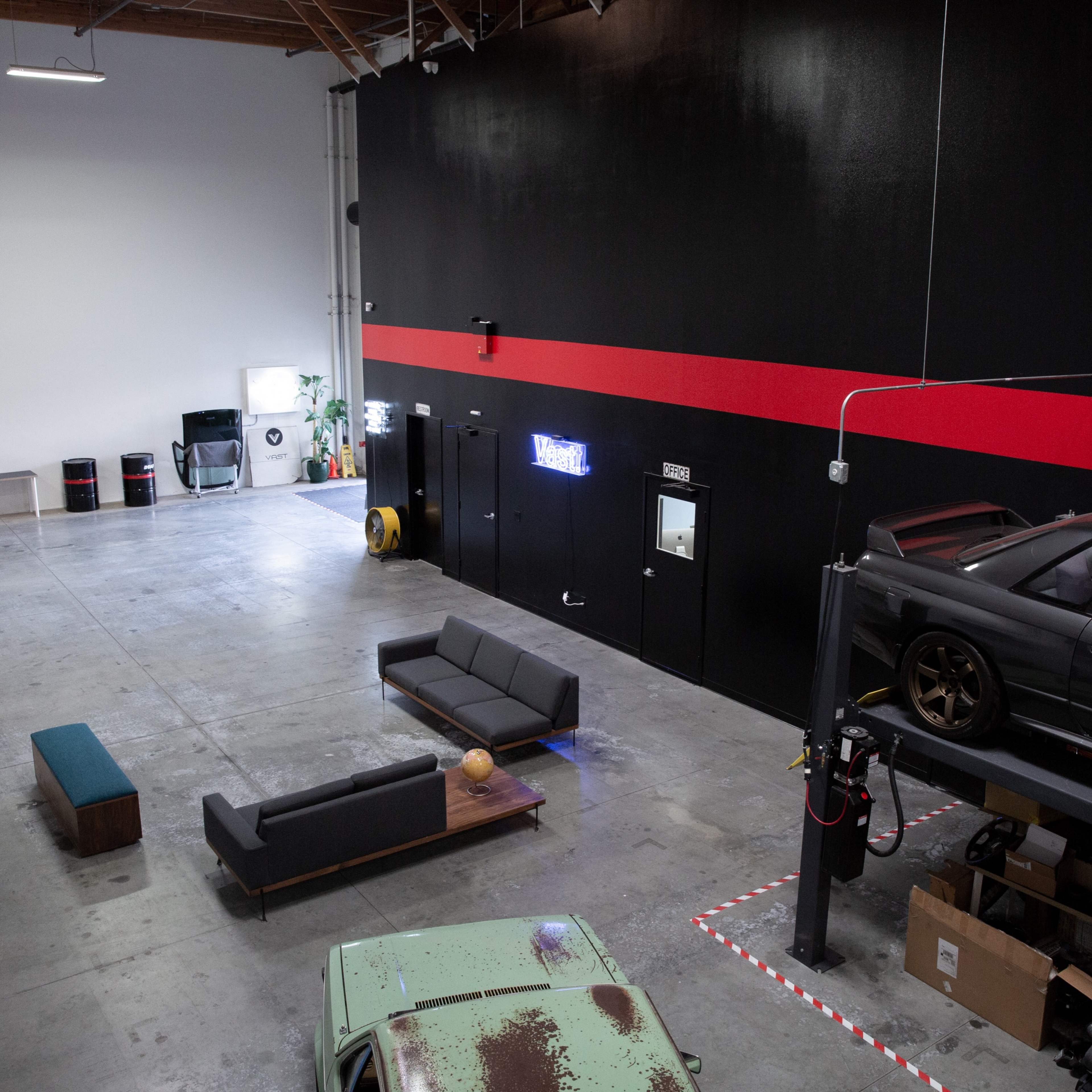 A spacious, unfurnished interior of a garage or warehouse with a gray concrete floor, a large black wall featuring a red stripe, a car on a lift, and some minimal furniture.