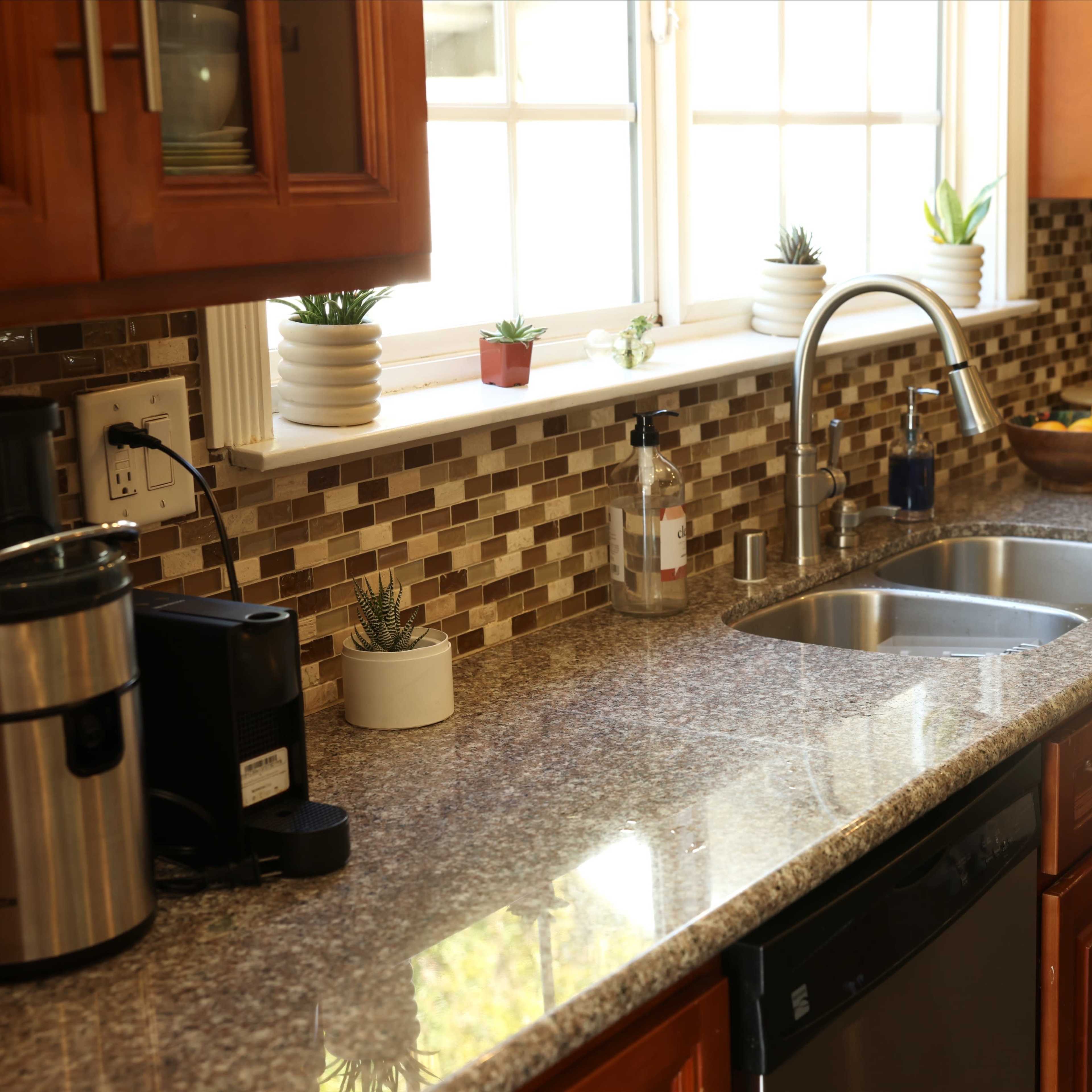 A kitchen with brown cabinets, a double sink, and a countertop adorned with small plants and a bowl of fruit, featuring a tiled backsplash.