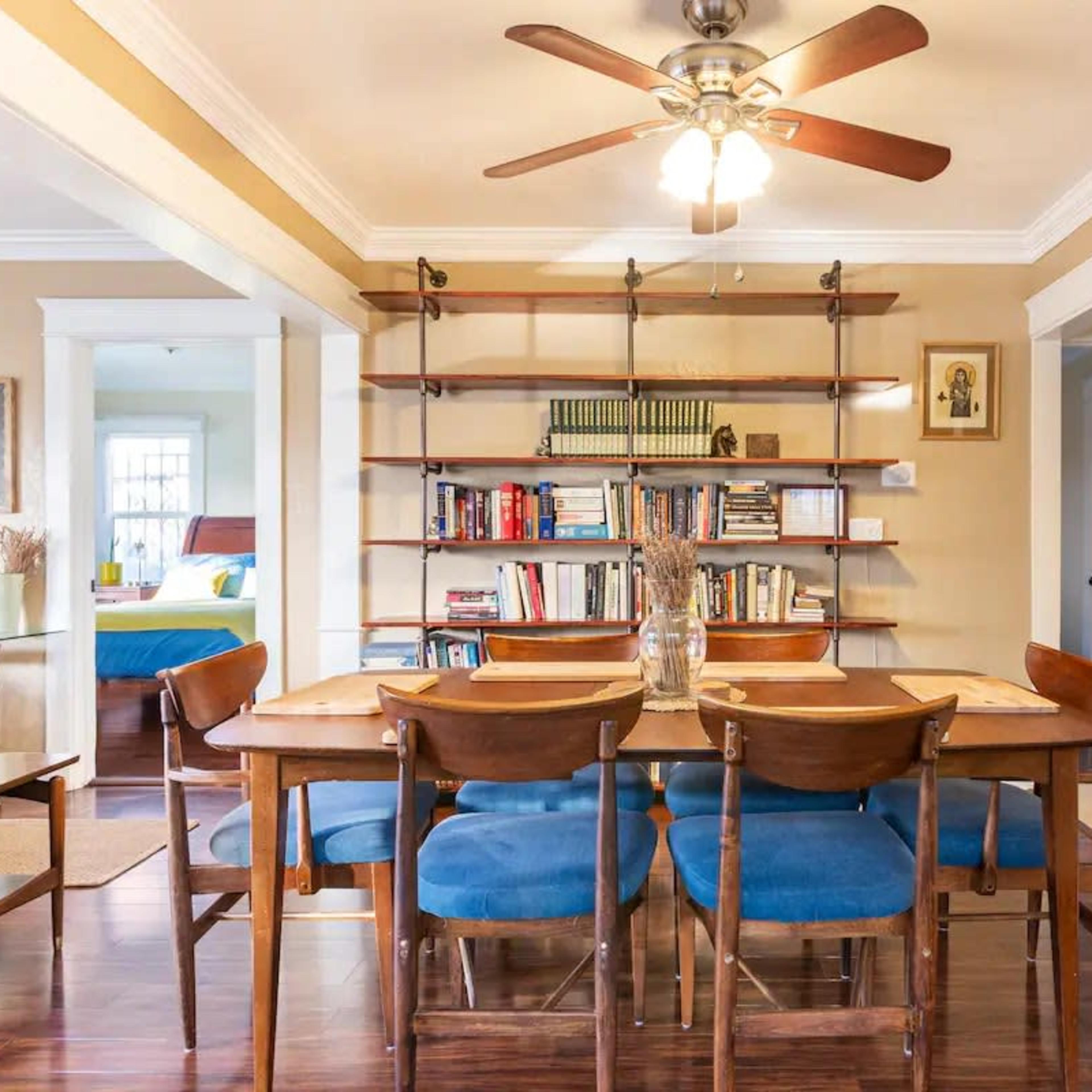A dining area with a wooden table surrounded by blue chairs, a ceiling fan above, and a bookshelf filled with books in the background.