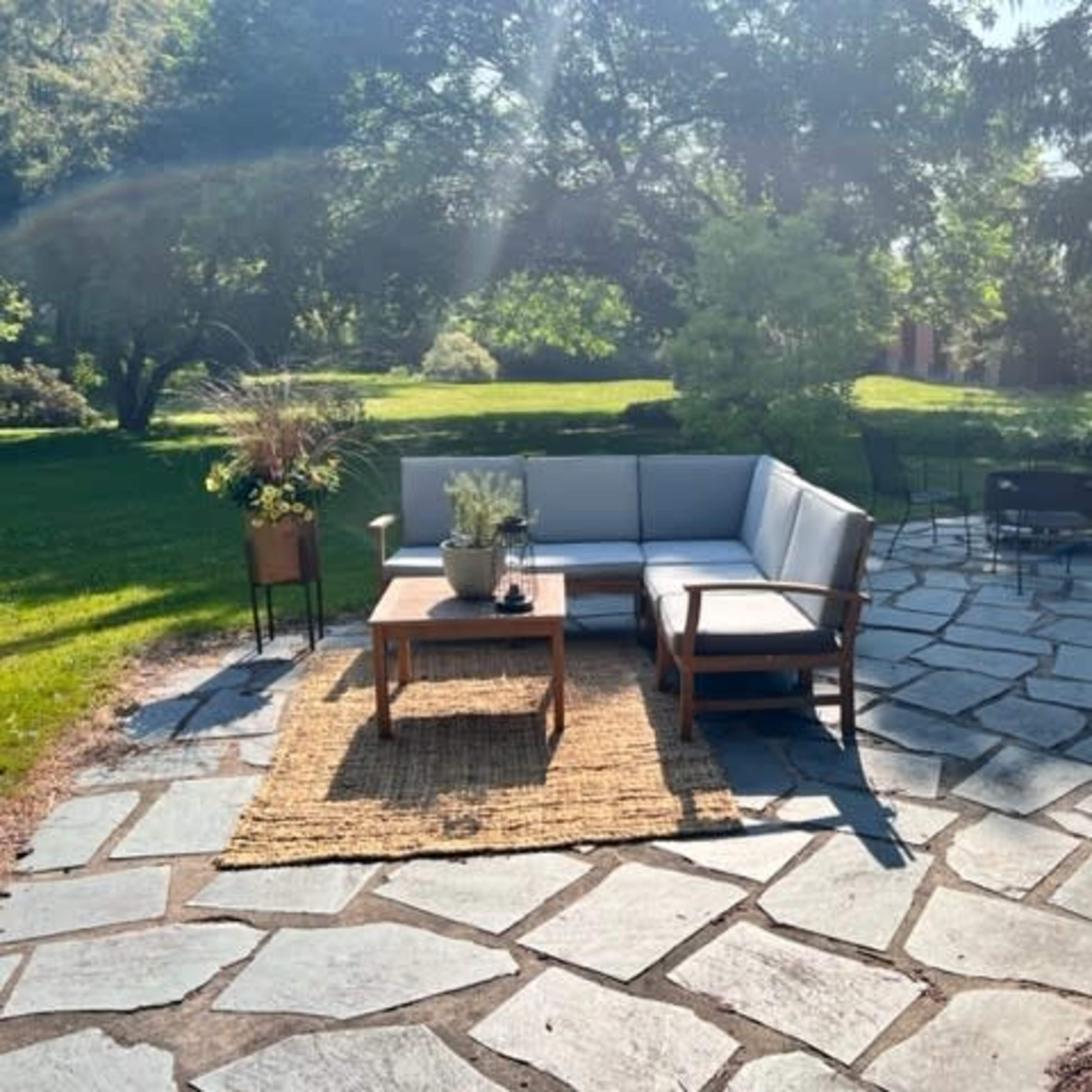 A patio area features a sectional sofa, a wooden coffee table, and a woven rug, surrounded by a stone pathway and various plants.