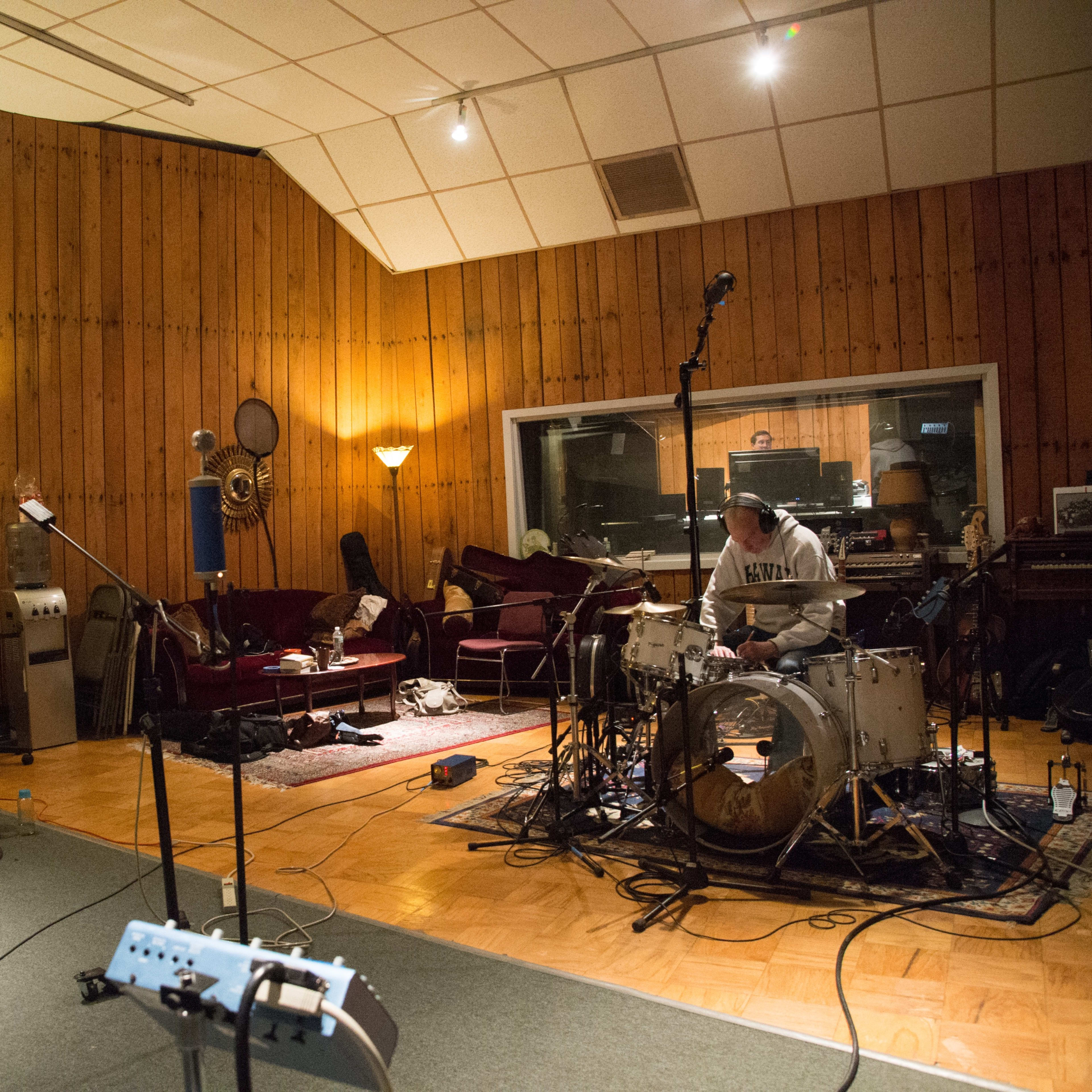 A music recording studio features a drummer on a set surrounded by cables and equipment, while a pianist sits nearby with a device in hand.