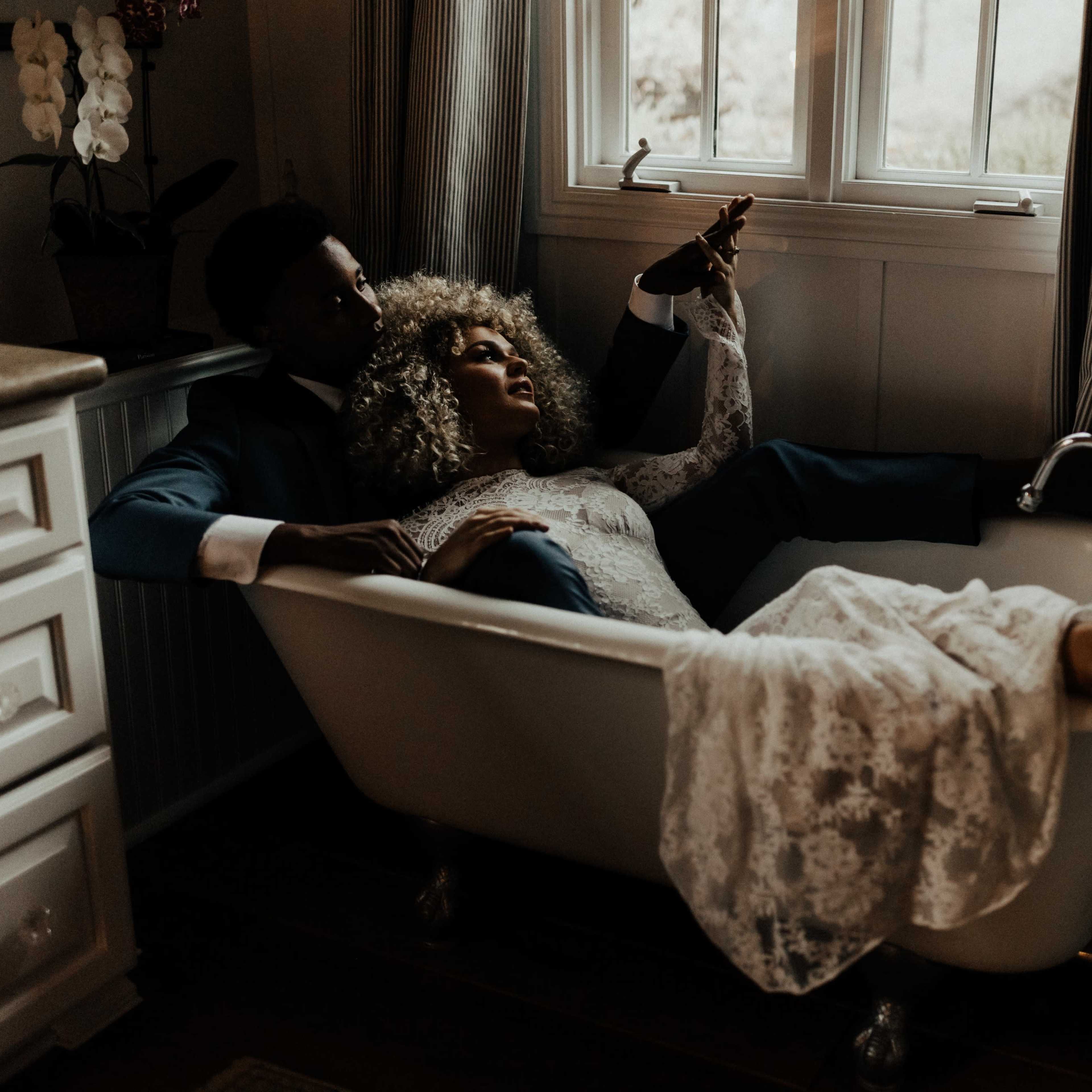 A couple sits together in a vintage bathtub, dressed elegantly, with the woman reclining against the man while reaching for something outside the frame.
