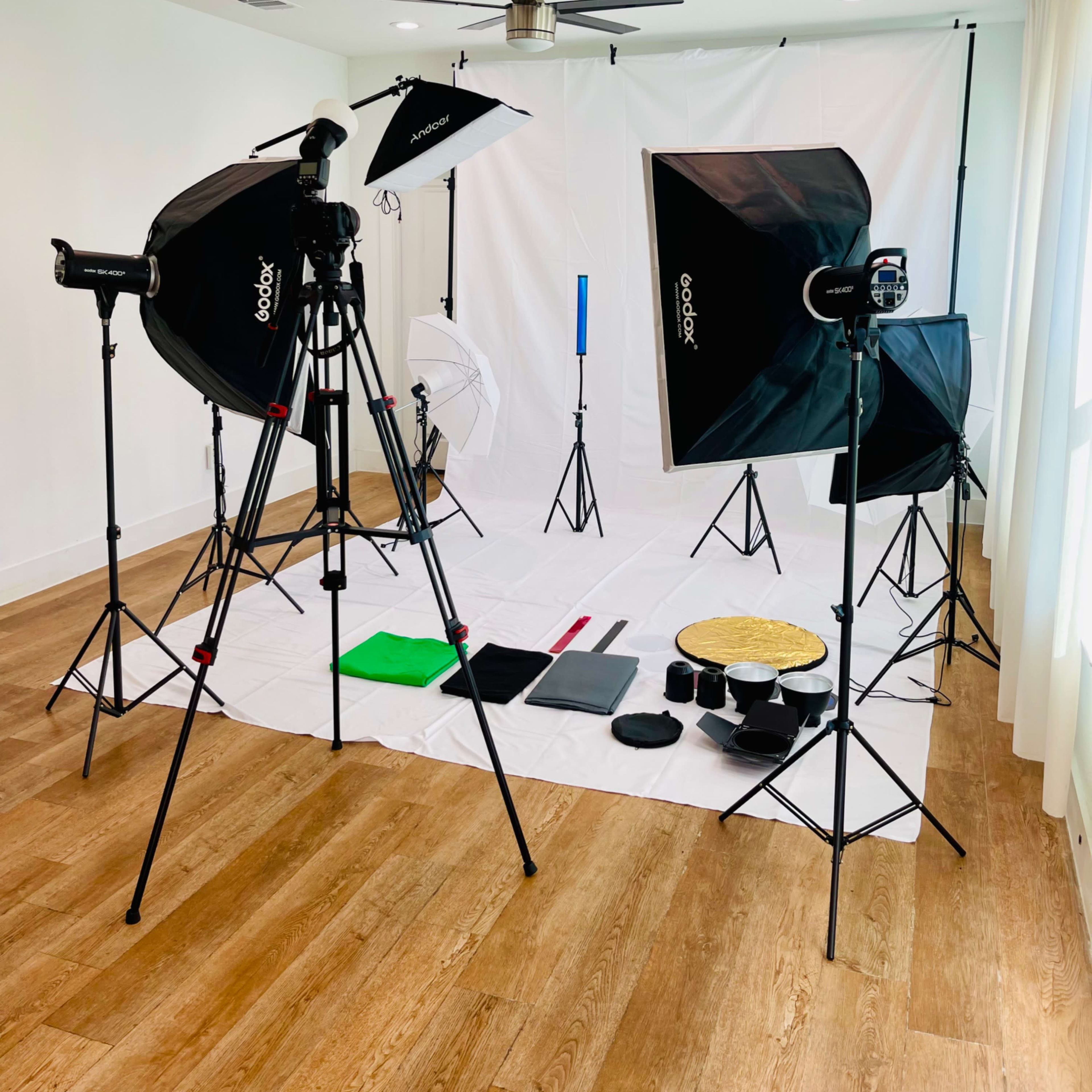A photography studio setup features softbox lights, tripods, and a white backdrop with various colored props arranged on the floor.