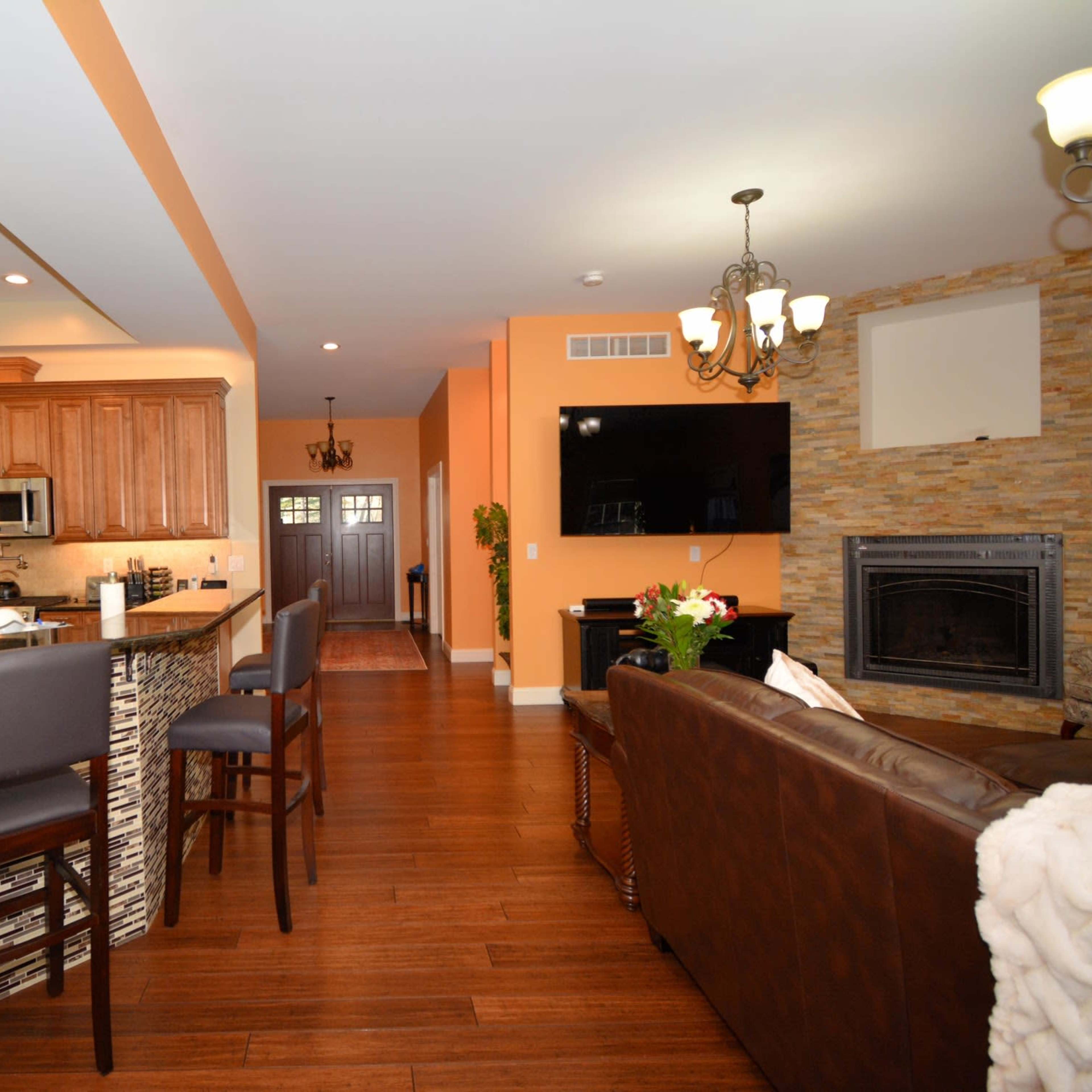 A well-lit open-concept living space featuring a kitchen with wooden cabinetry, a dining area, and a living room with a stone fireplace and large TV.