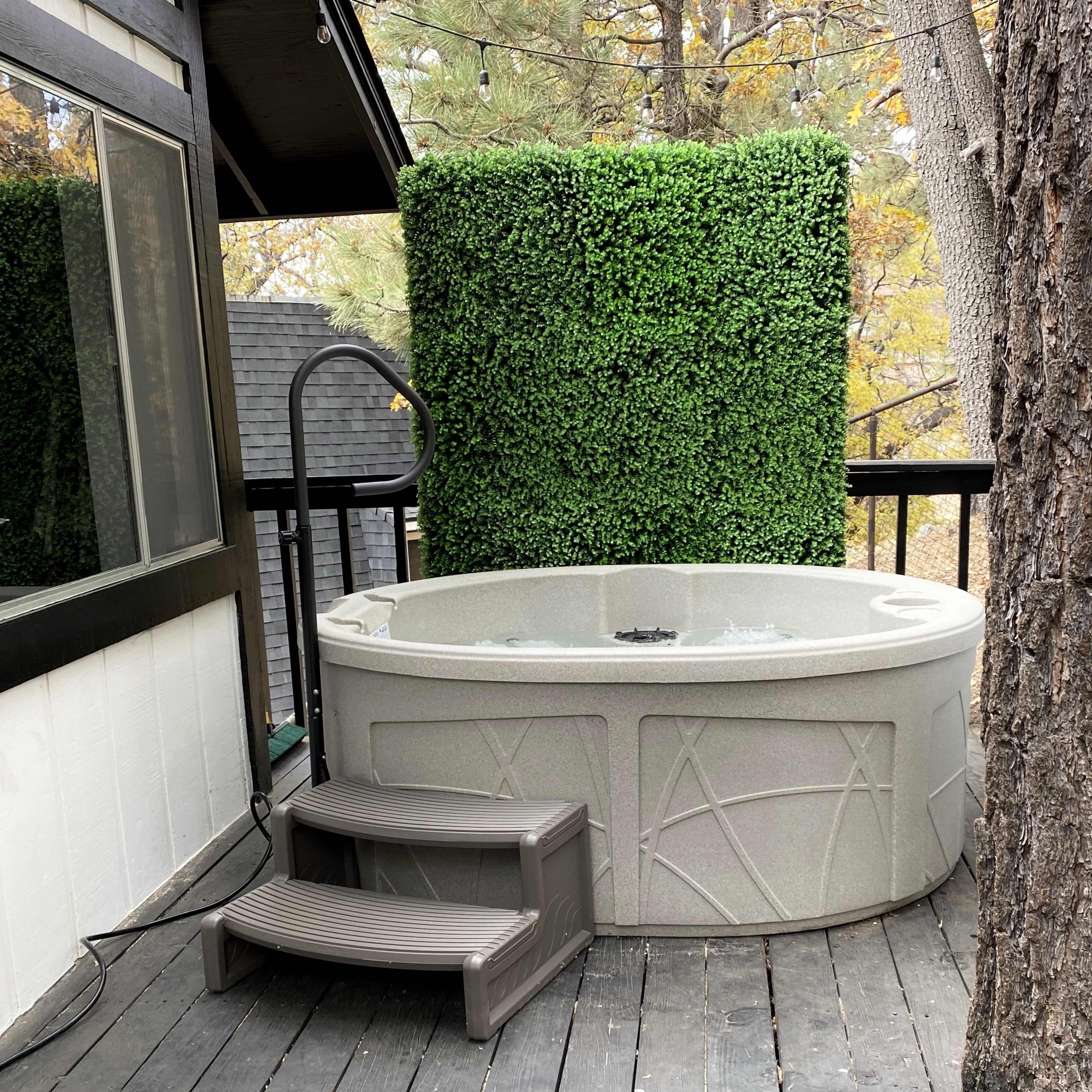 A hot tub is situated on a wooden deck next to a house, surrounded by trees and a tall green hedge.