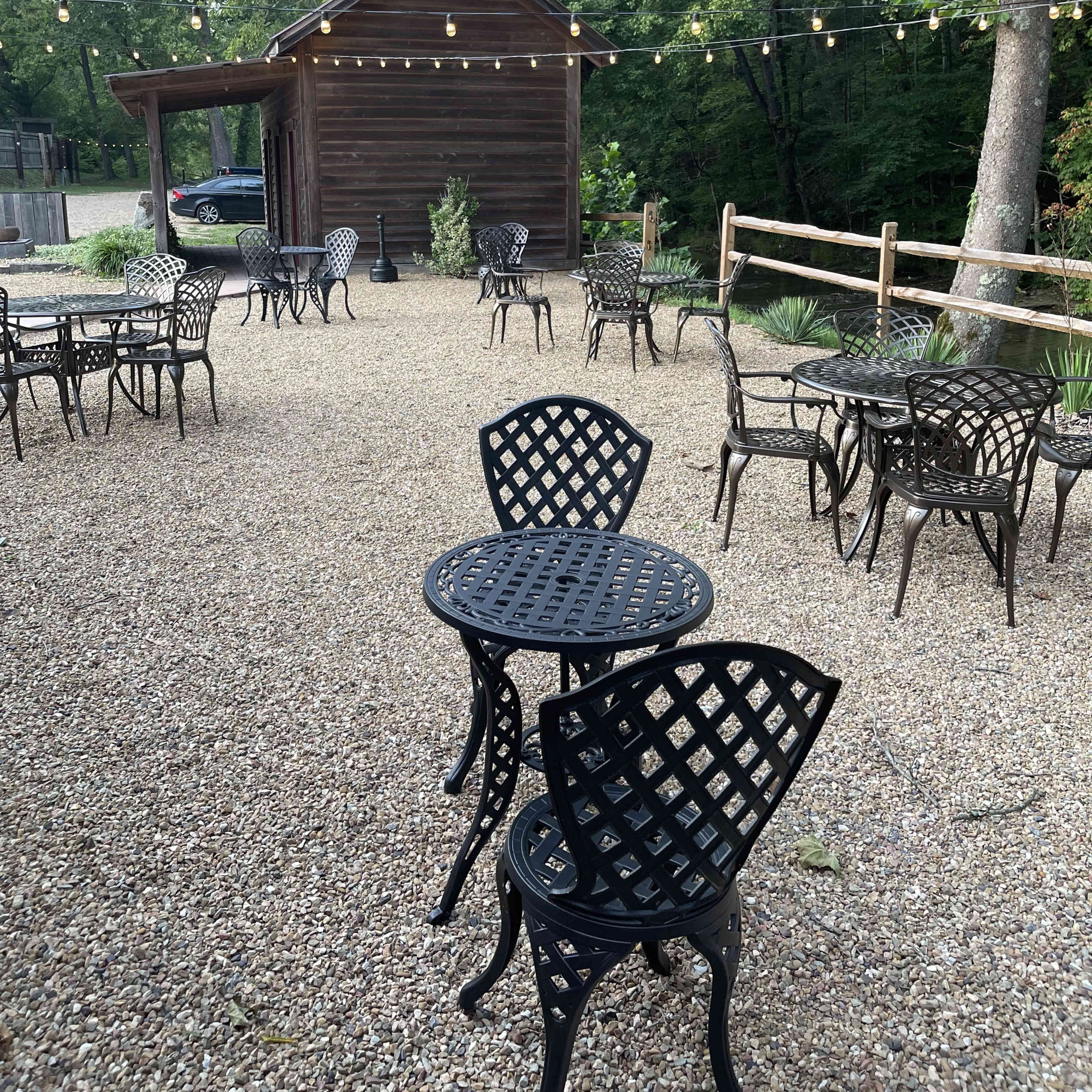 A gravel outdoor seating area features several black metal tables and chairs arranged around a rustic wooden cabin, with string lights overhead.