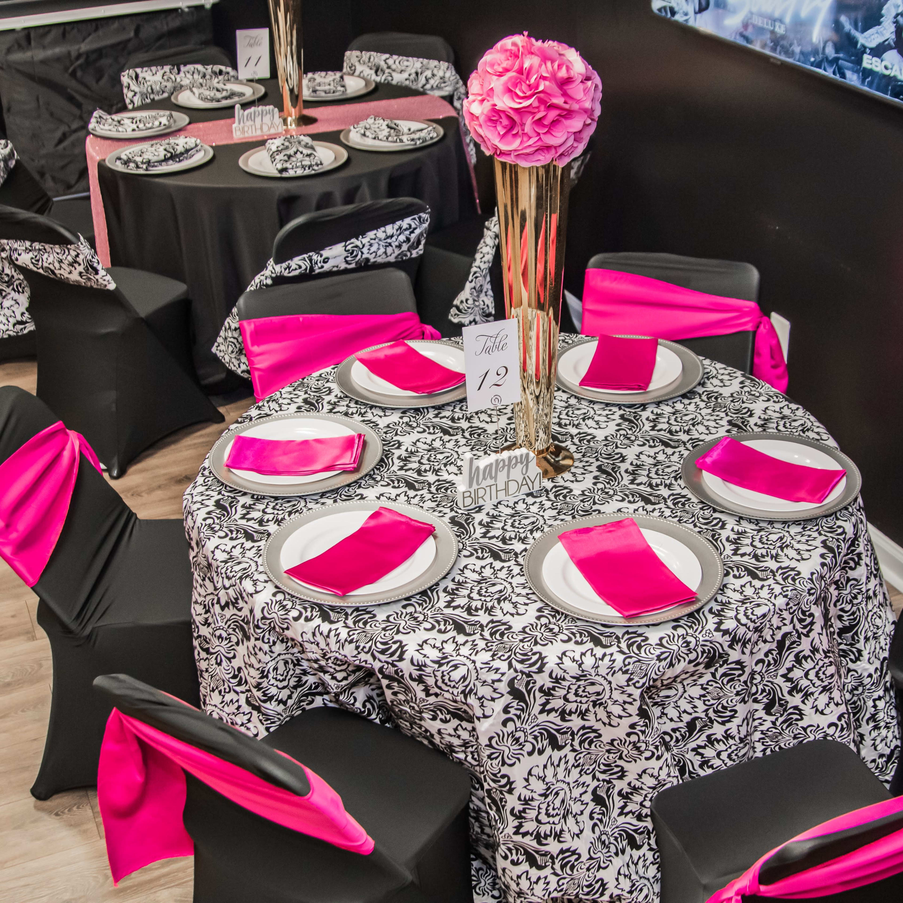 A dining area is set up with black and white patterned tablecloths, pink napkins, and tall centerpieces on each table.