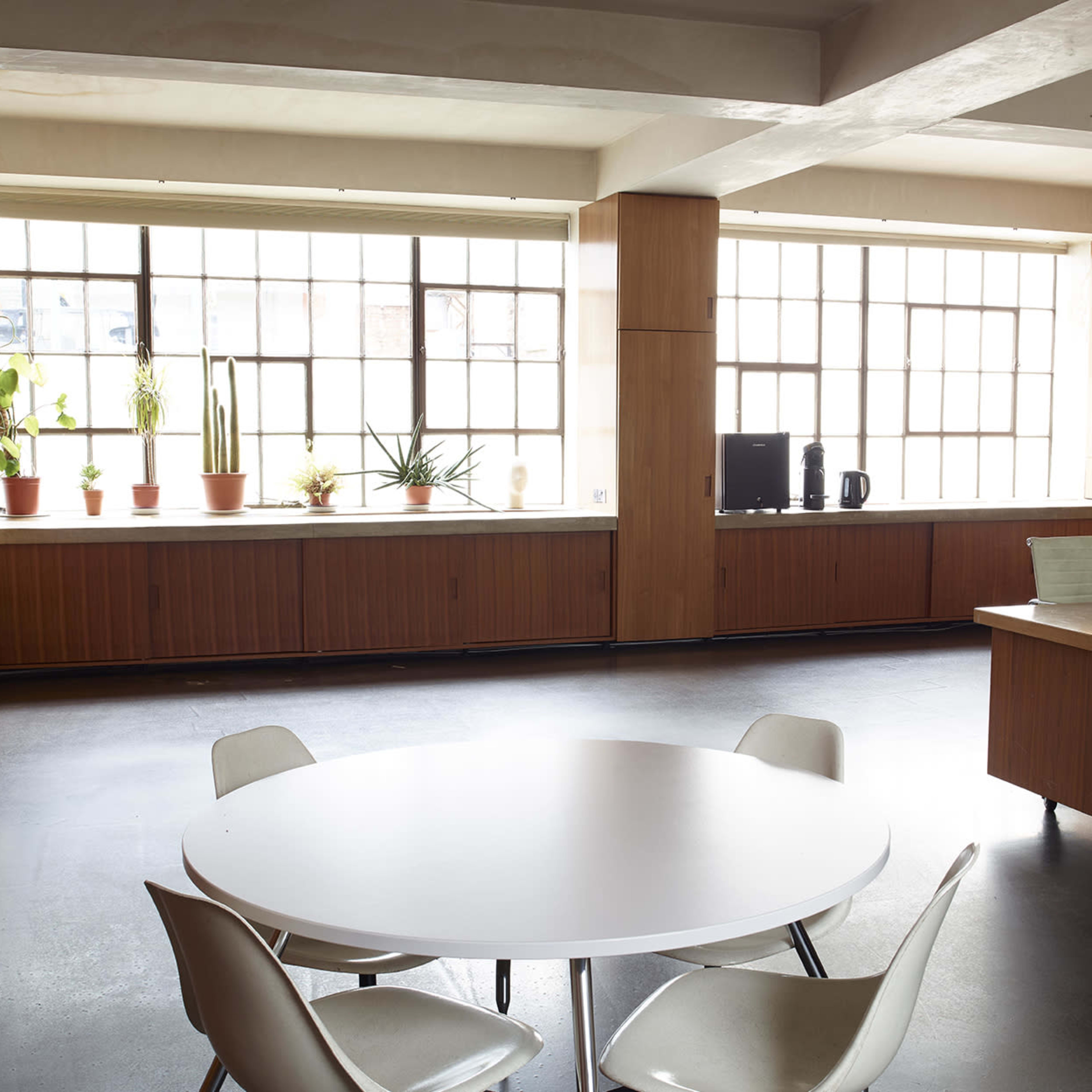 A spacious room featuring a circular table with four chairs, a brown sofa, and large windows lined with potted plants.