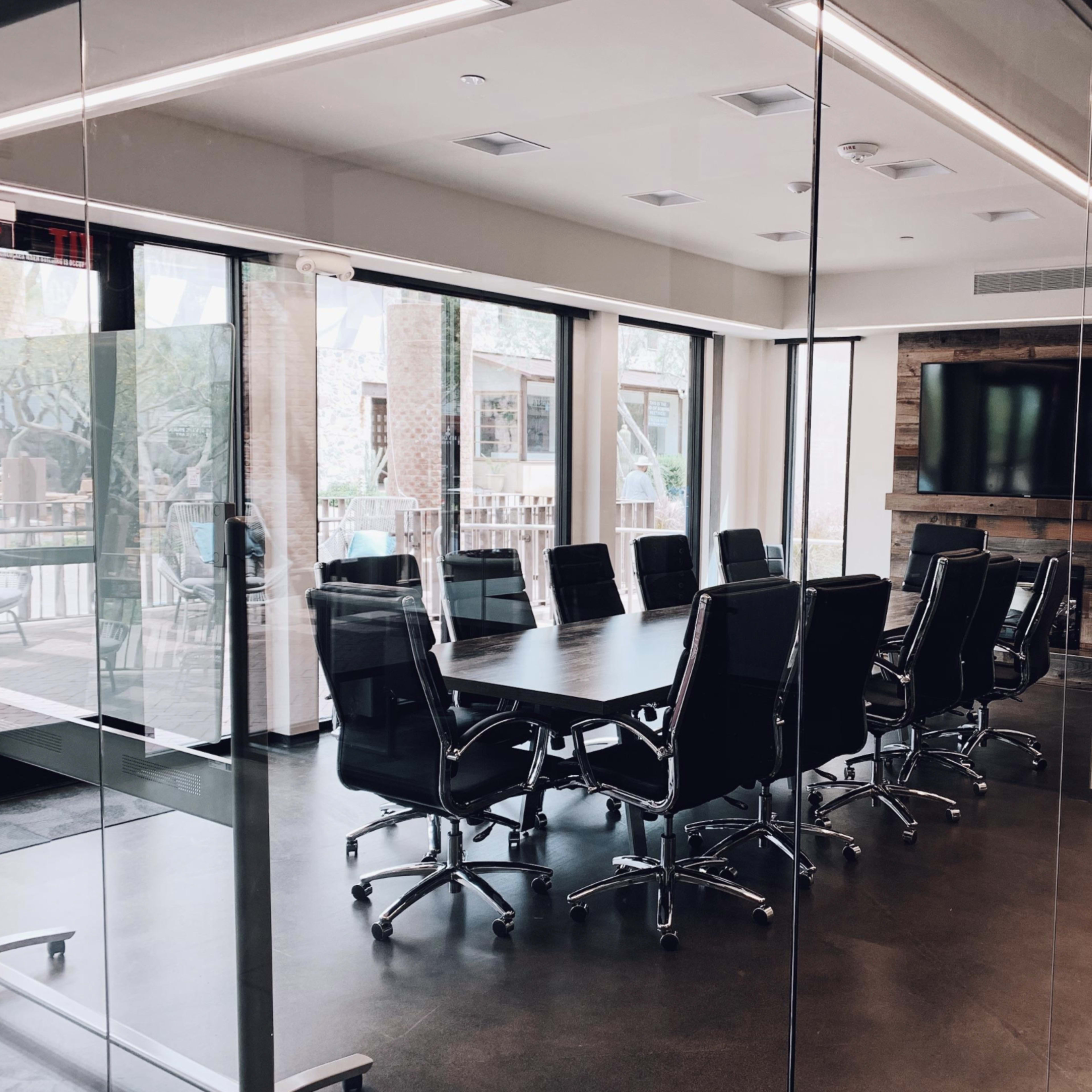 A large conference room features a long table surrounded by black leather chairs, with walls of glass providing a view of the outside.