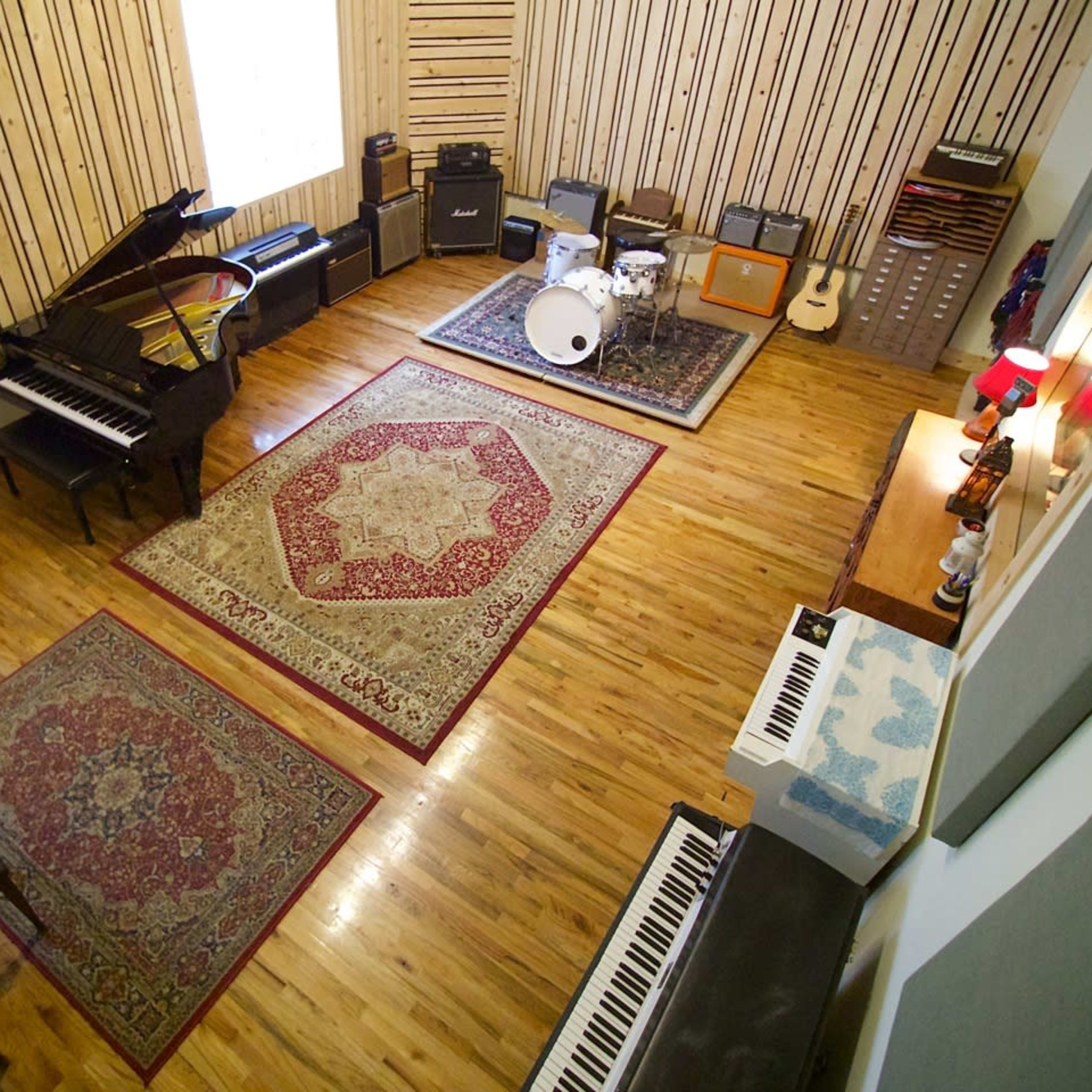 A spacious music studio featuring a grand piano, various amplifiers, a drum set, and several keyboards arranged on a hardwood floor with decorative rugs.