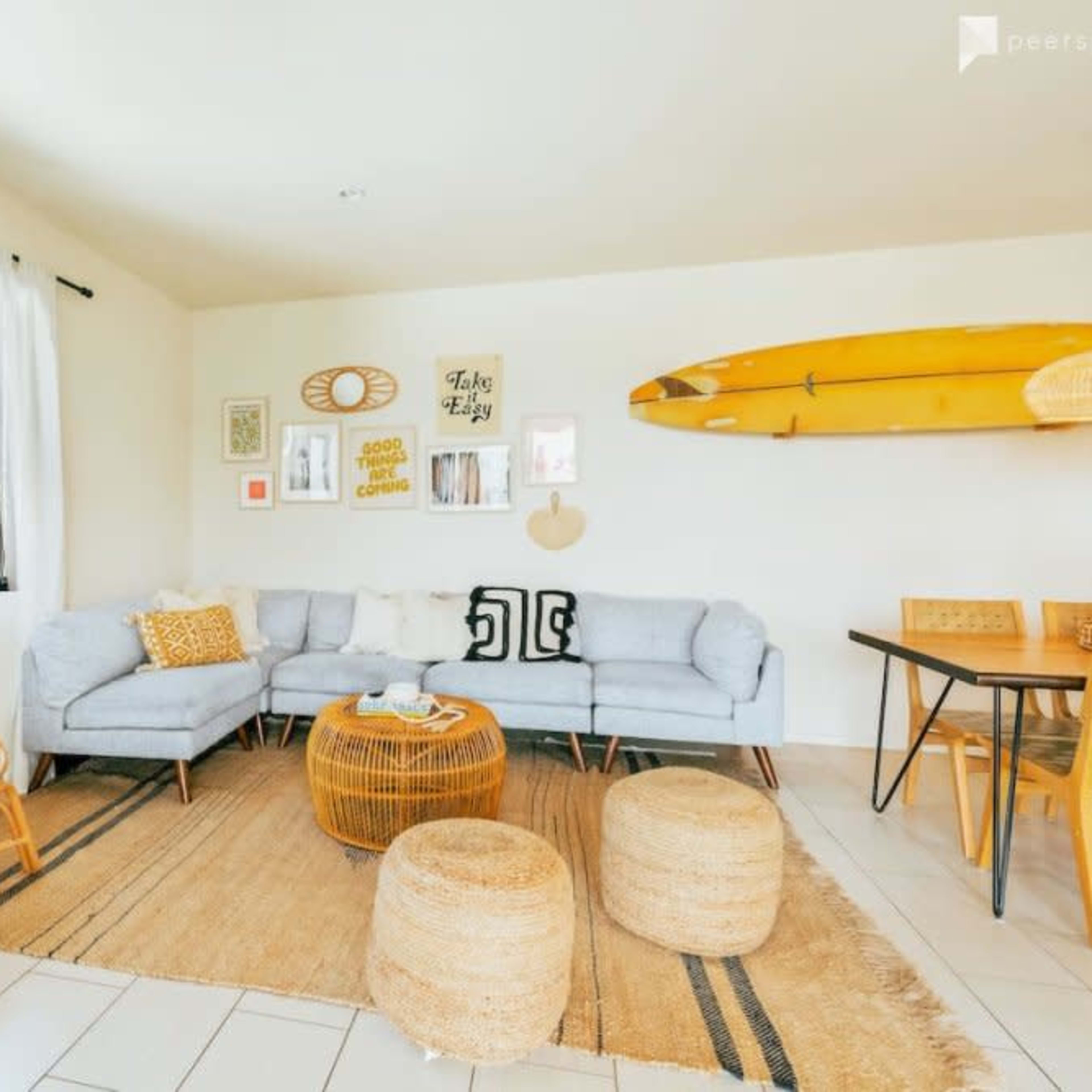 A modern living room features a sectional sofa, a round coffee table, woven stools, and a surfboard mounted on the wall.