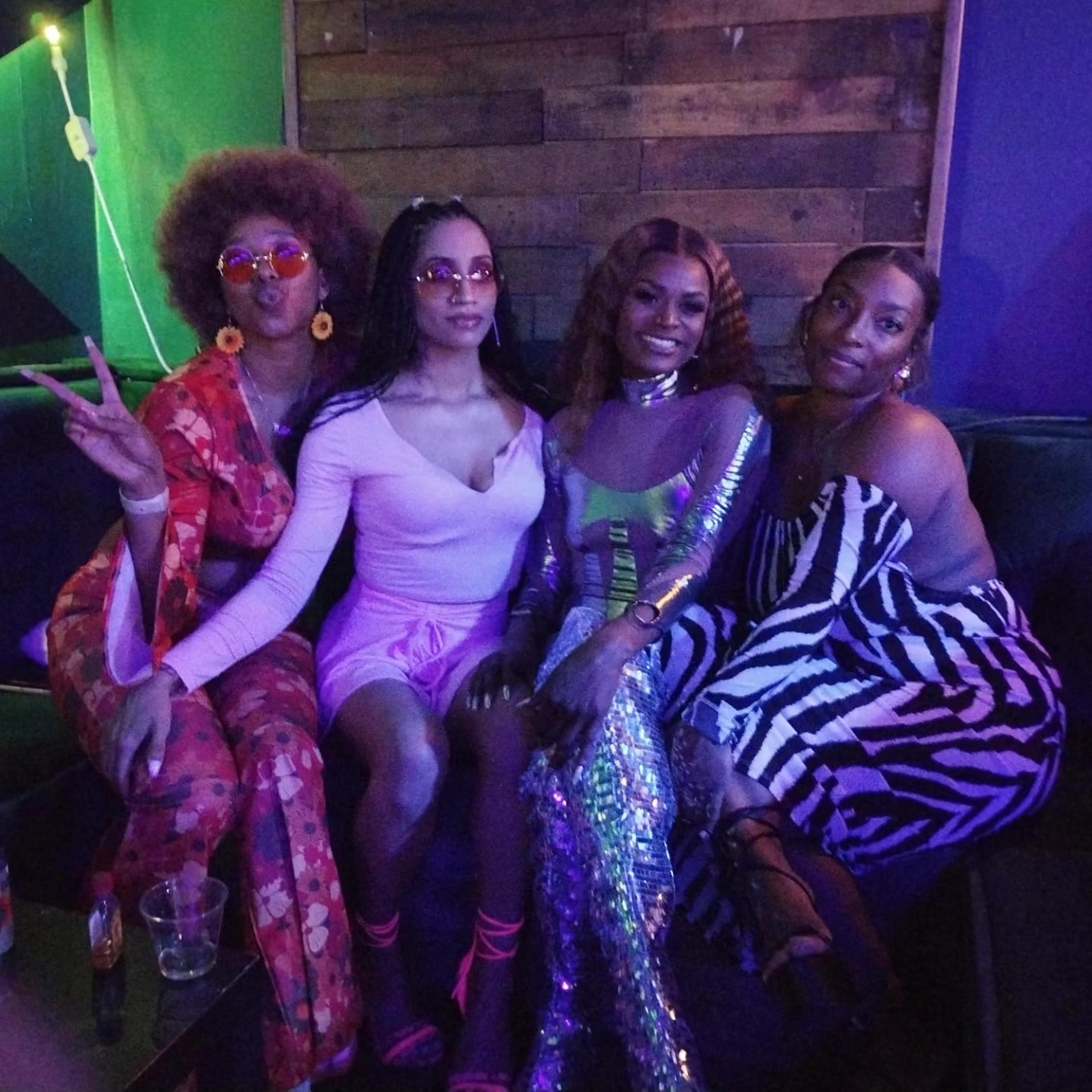Four women pose together on a couch in a dimly lit venue, each wearing distinct outfits and accessories.