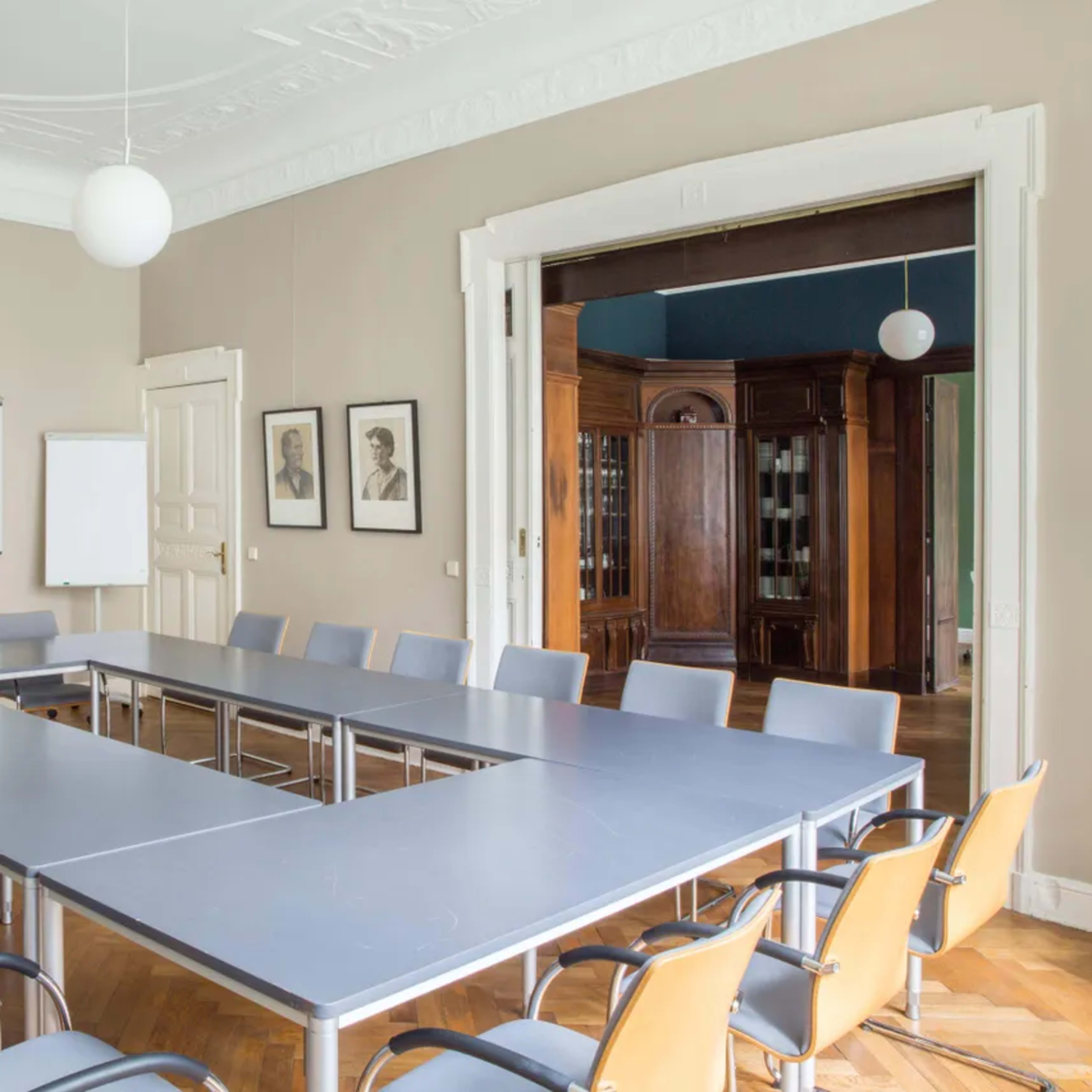A conference room with a large rectangular table surrounded by chairs, along with whiteboards and framed portraits on the walls.