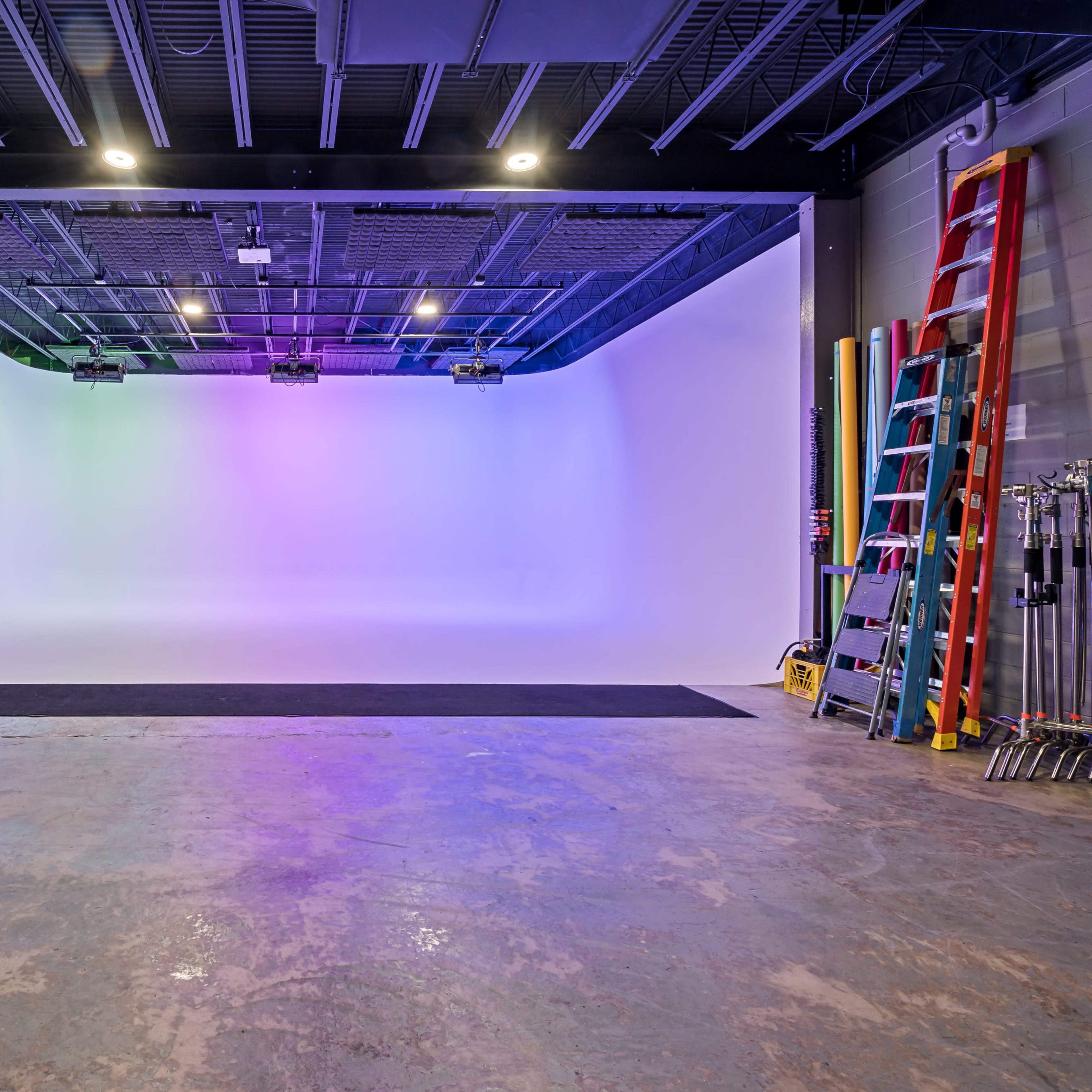 An empty photo studio with a curved backdrop, colorful lighting, and various equipment visible in the background.