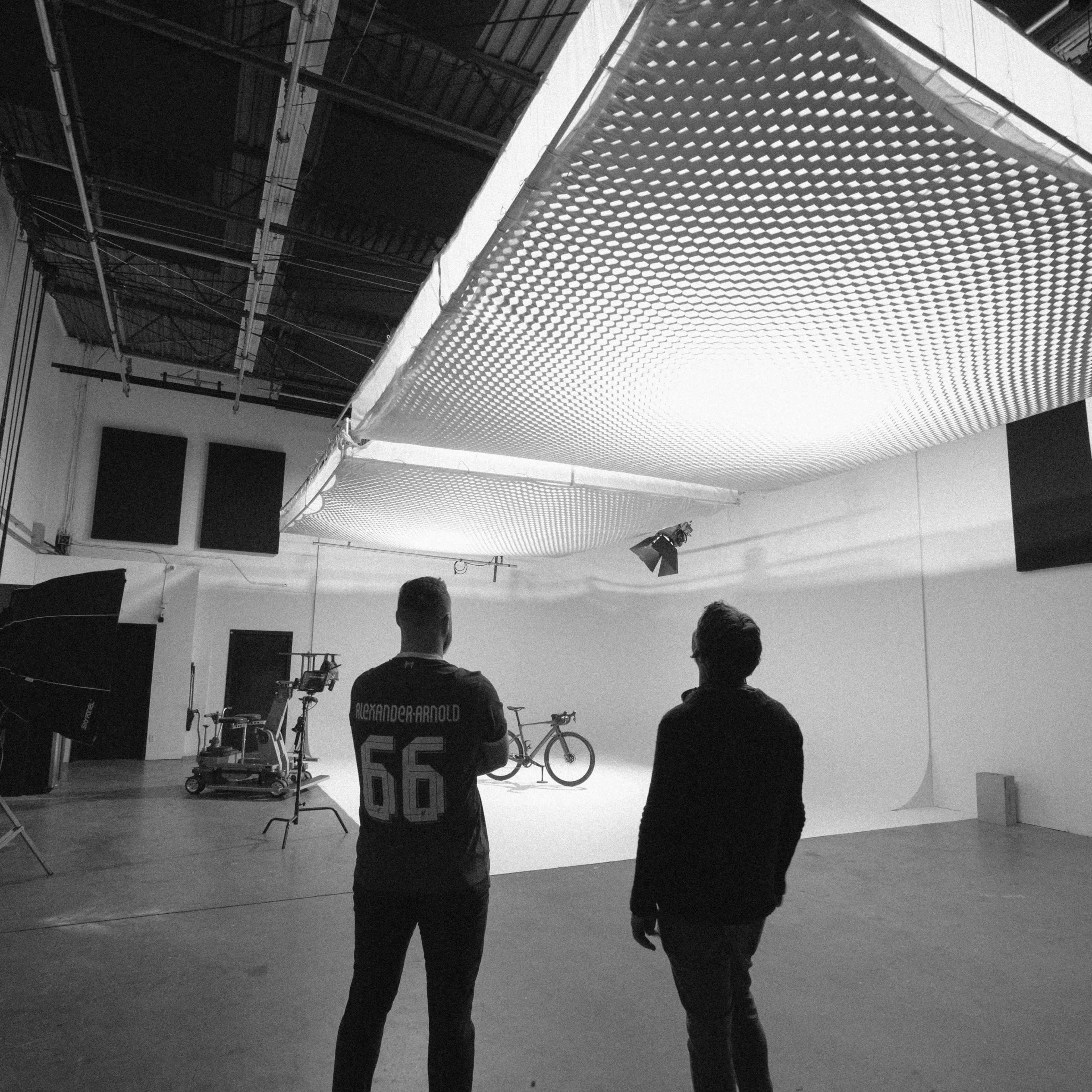 Two people stand in a photography studio, observing a large light diffusing panel suspended above a white backdrop.