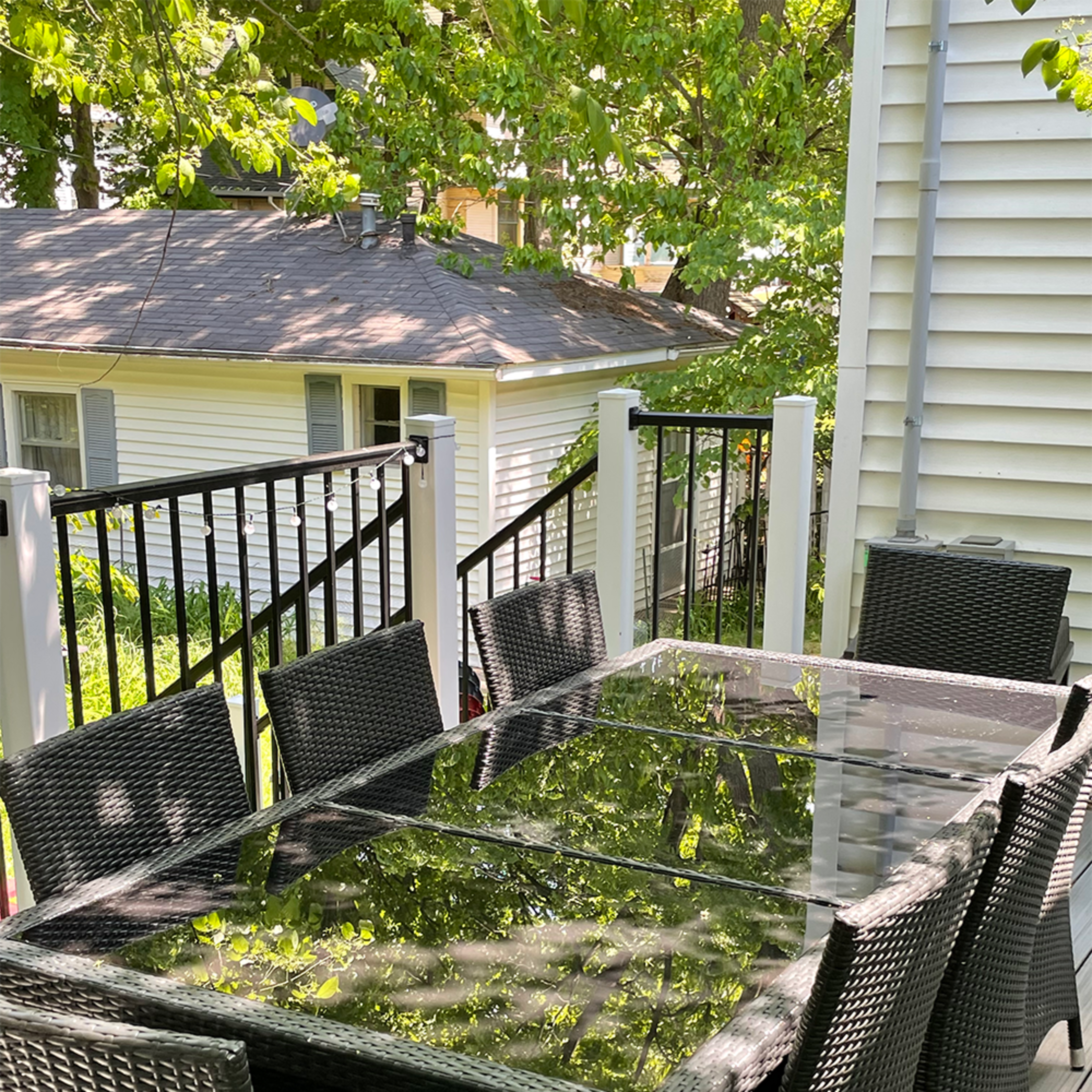A large glass-top outdoor dining table surrounded by black wicker chairs is positioned on a deck, with greenery reflected in the table's surface.