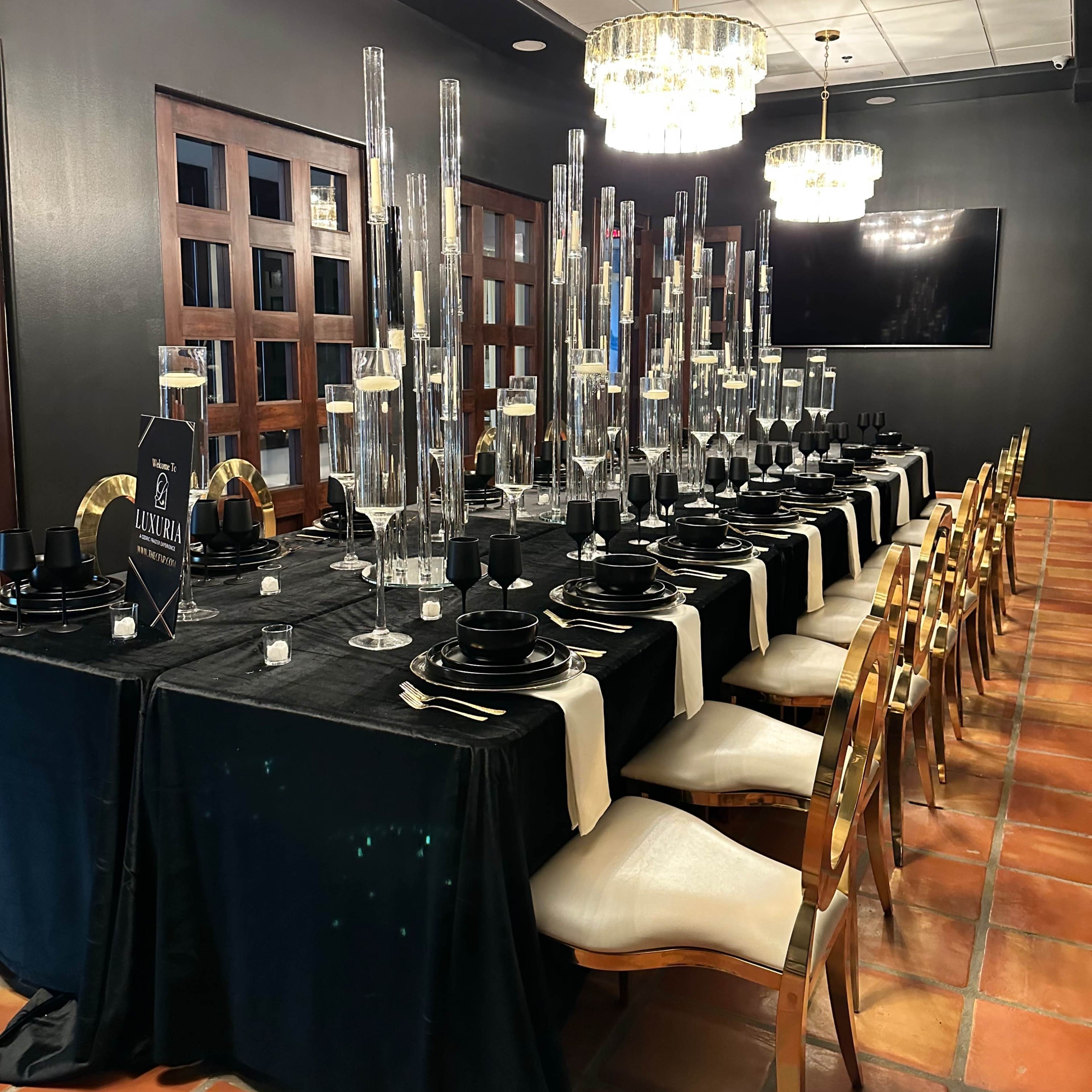 A long, elegantly set dining table with black and gold tableware, surrounded by tall candle holders, in a dimly lit room.