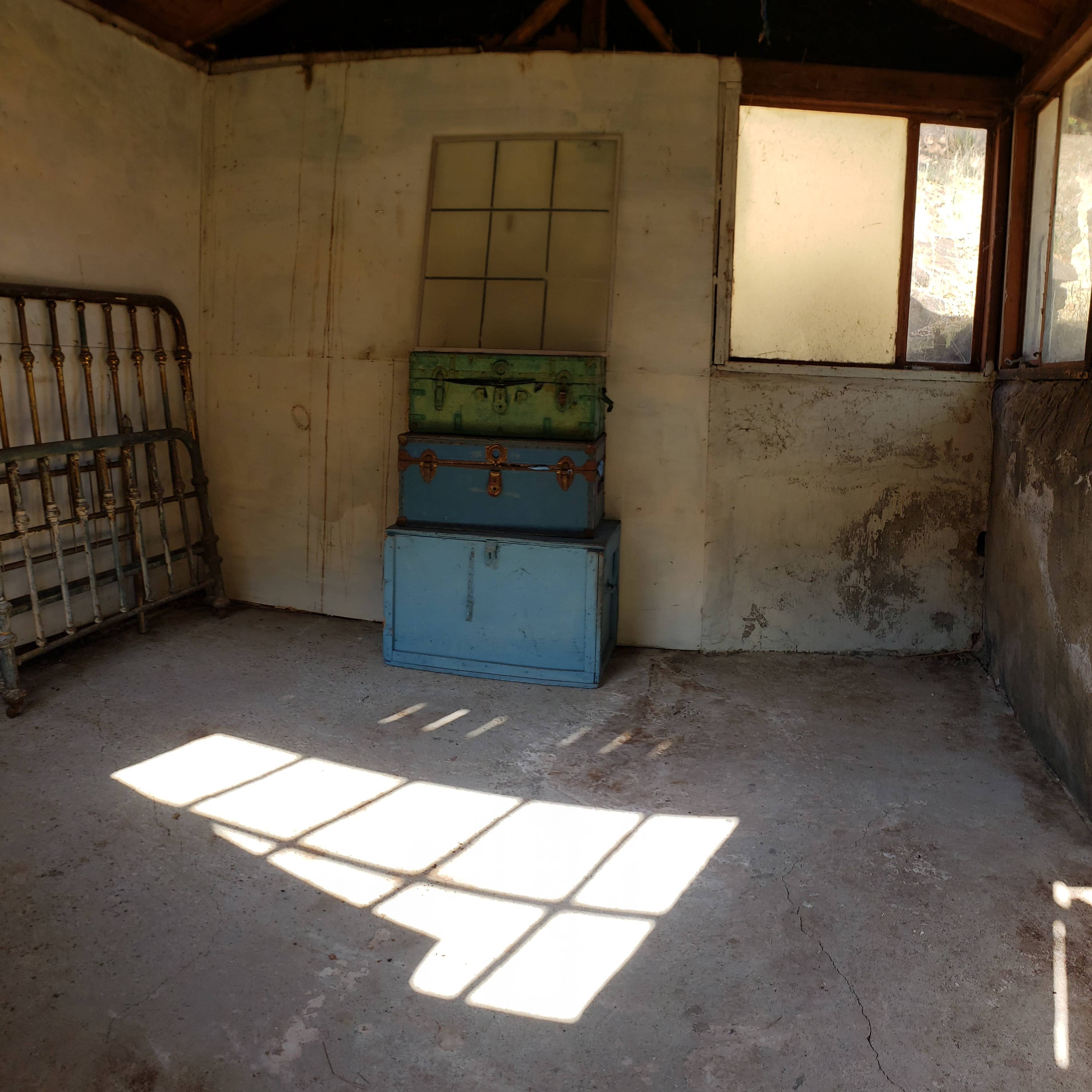 An empty, rustic room with a green and blue trunk stacked on the floor next to a wooden bed frame and a window casting shadows on the concrete floor.