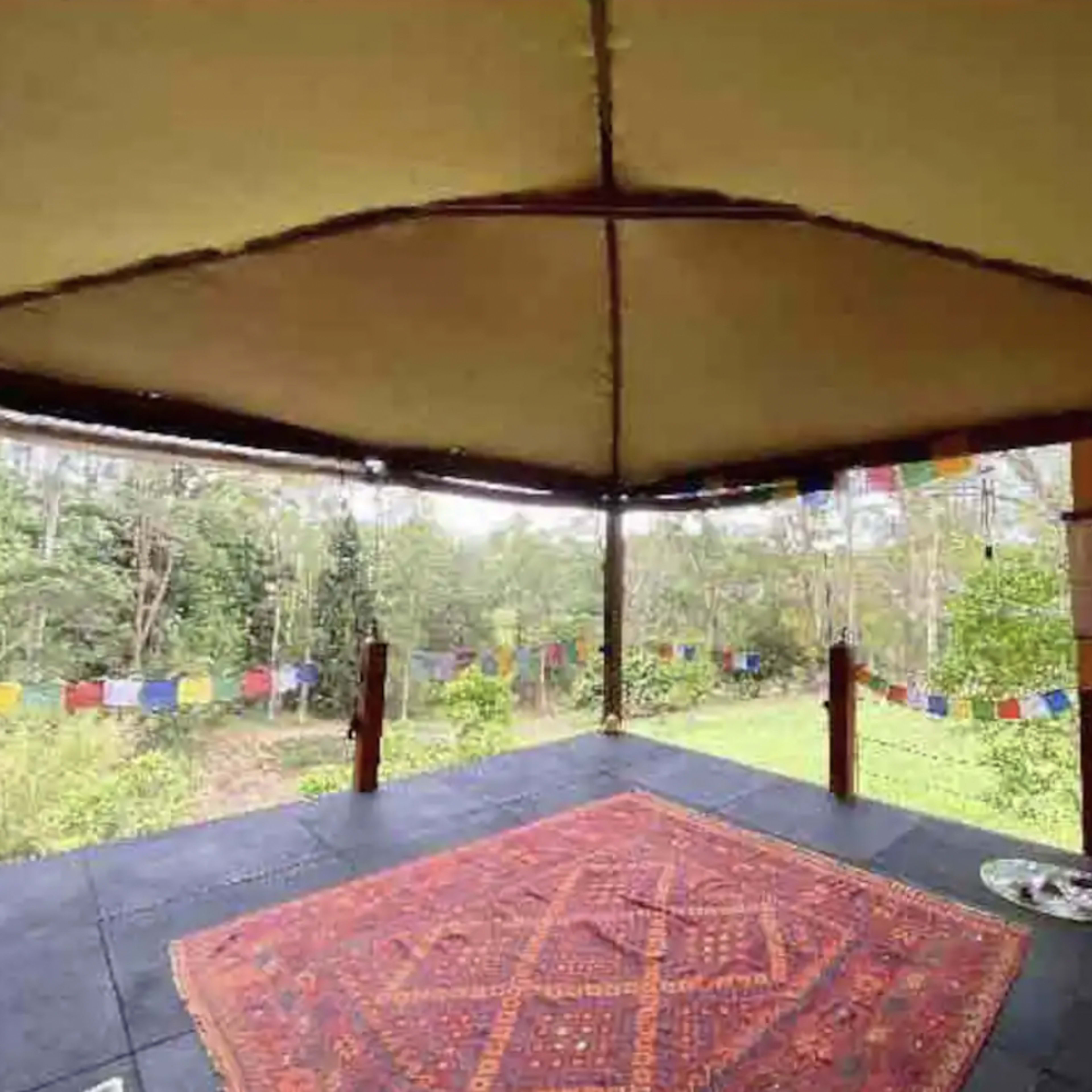 A spacious outdoor pavilion with a patterned rug and colorful prayer flags visible against a green backdrop.