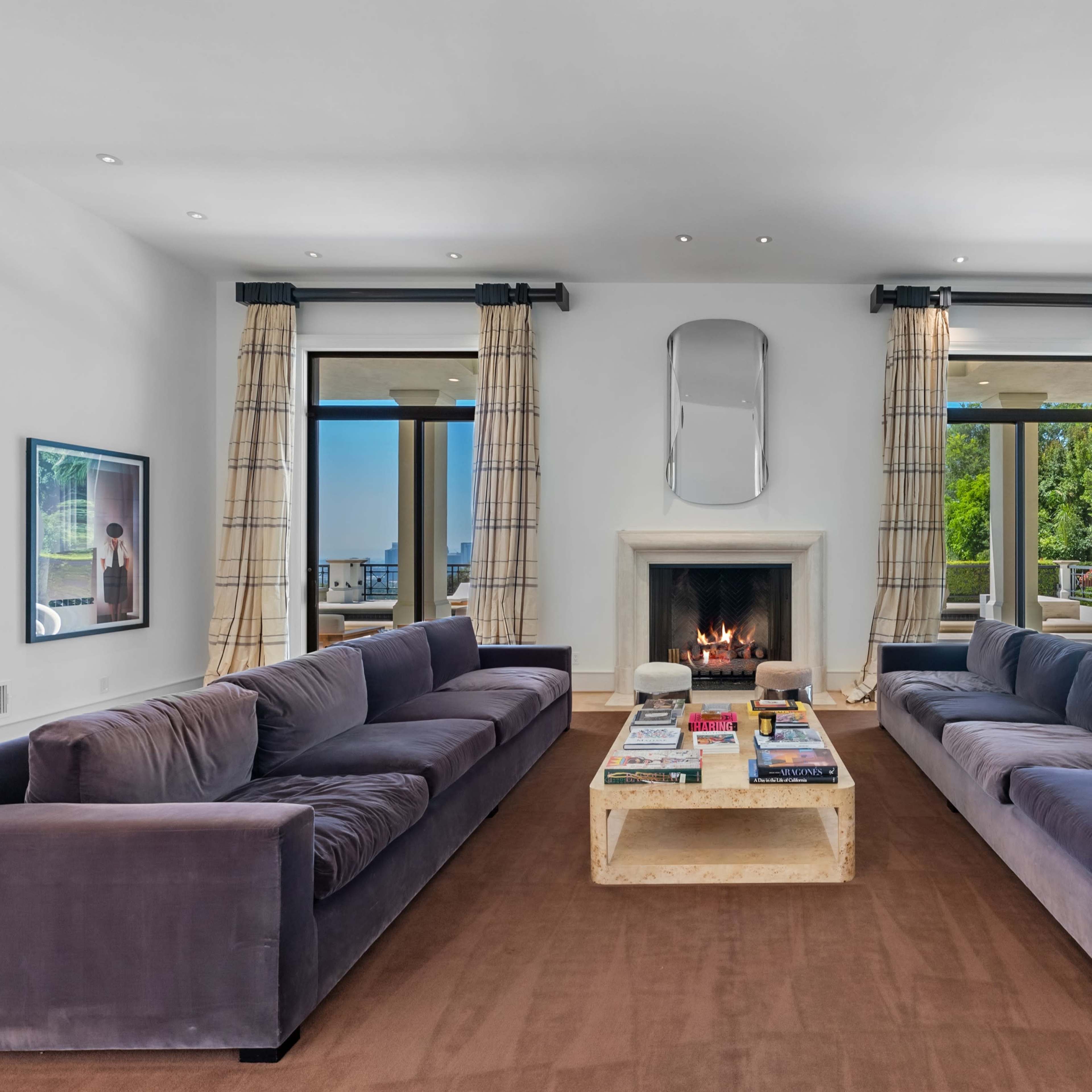 A modern living room with two large sofas facing a fireplace, framed by large windows and light-colored curtains.
