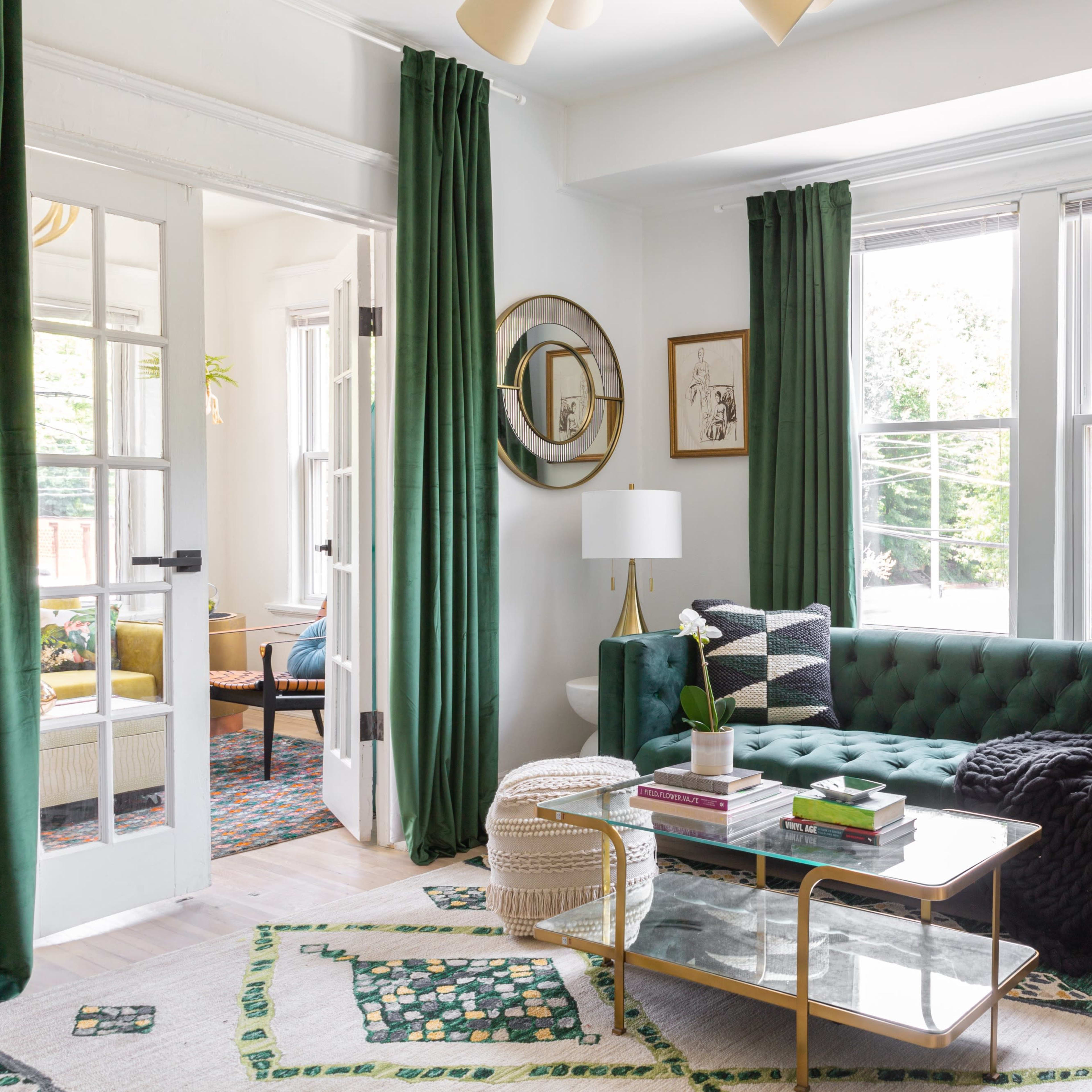 A bright living room features a green tufted sofa, a glass coffee table, and large windows adorned with green curtains.