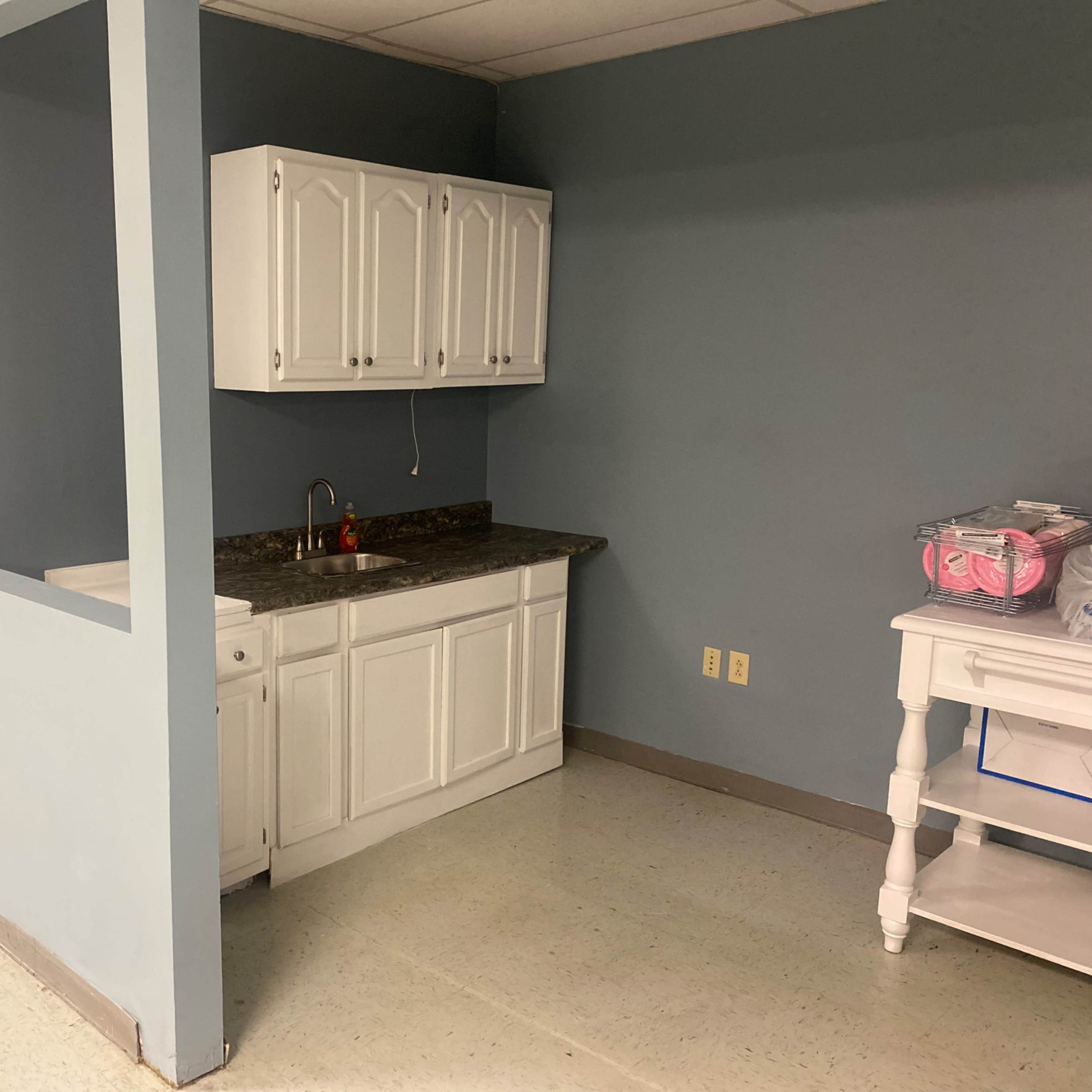 A small kitchenette with white cabinets and a sink, situated in a hallway with light-colored walls and flooring.