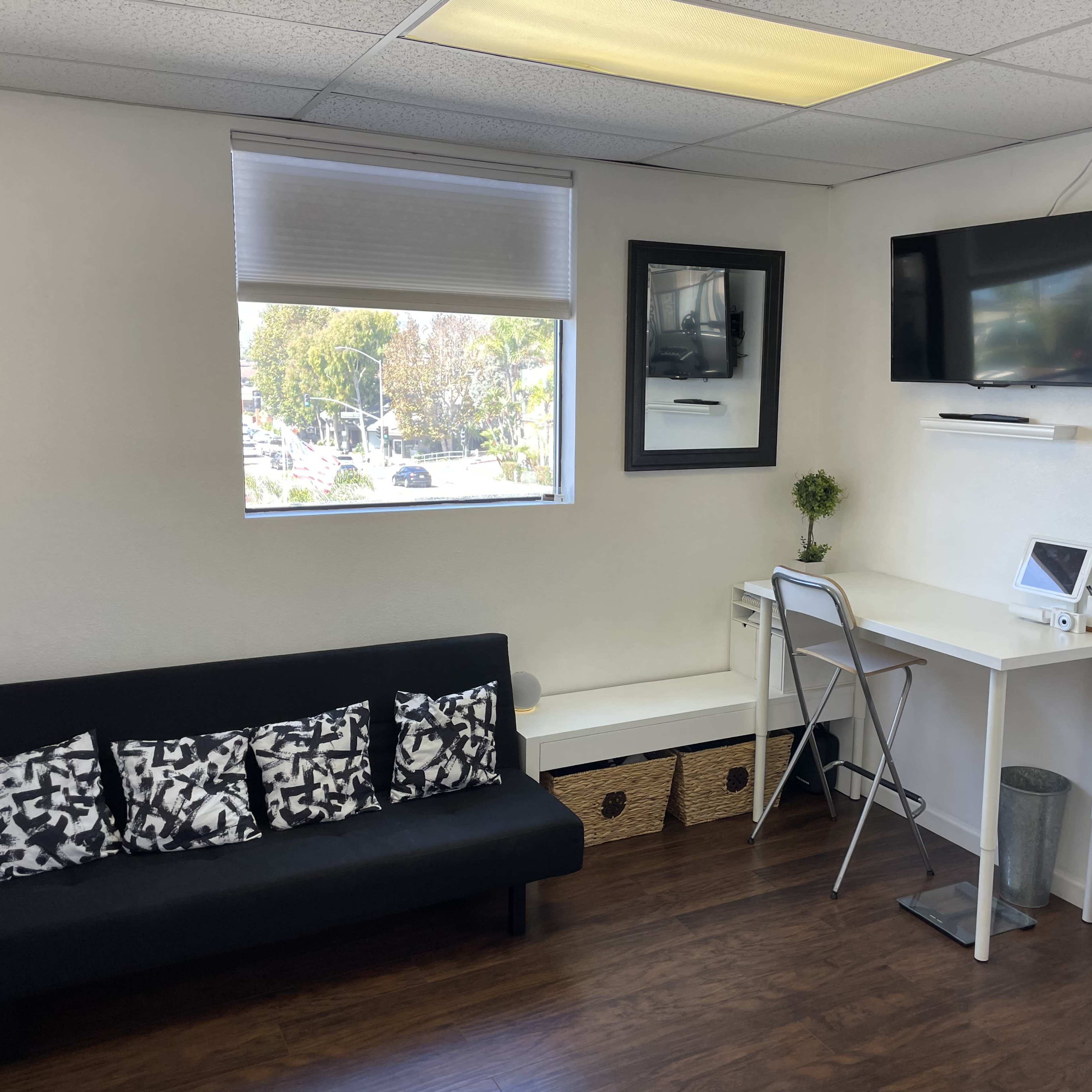 A small, modern office space with a black couch, a desk with a monitor, and a window overlooking a street.