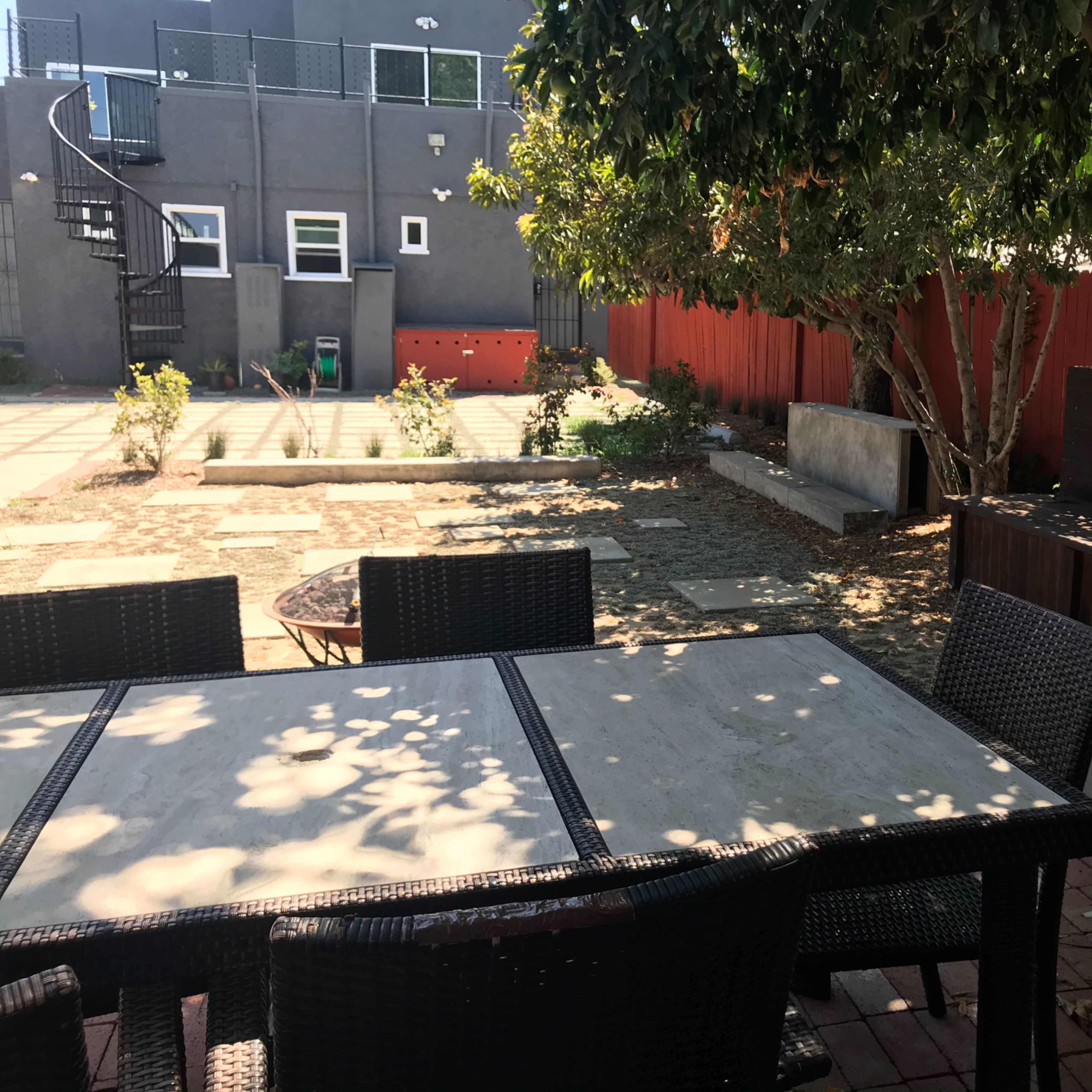 A landscaped outdoor area featuring a table and chairs, a spiral staircase in the background, and various plants and seating arrangements.