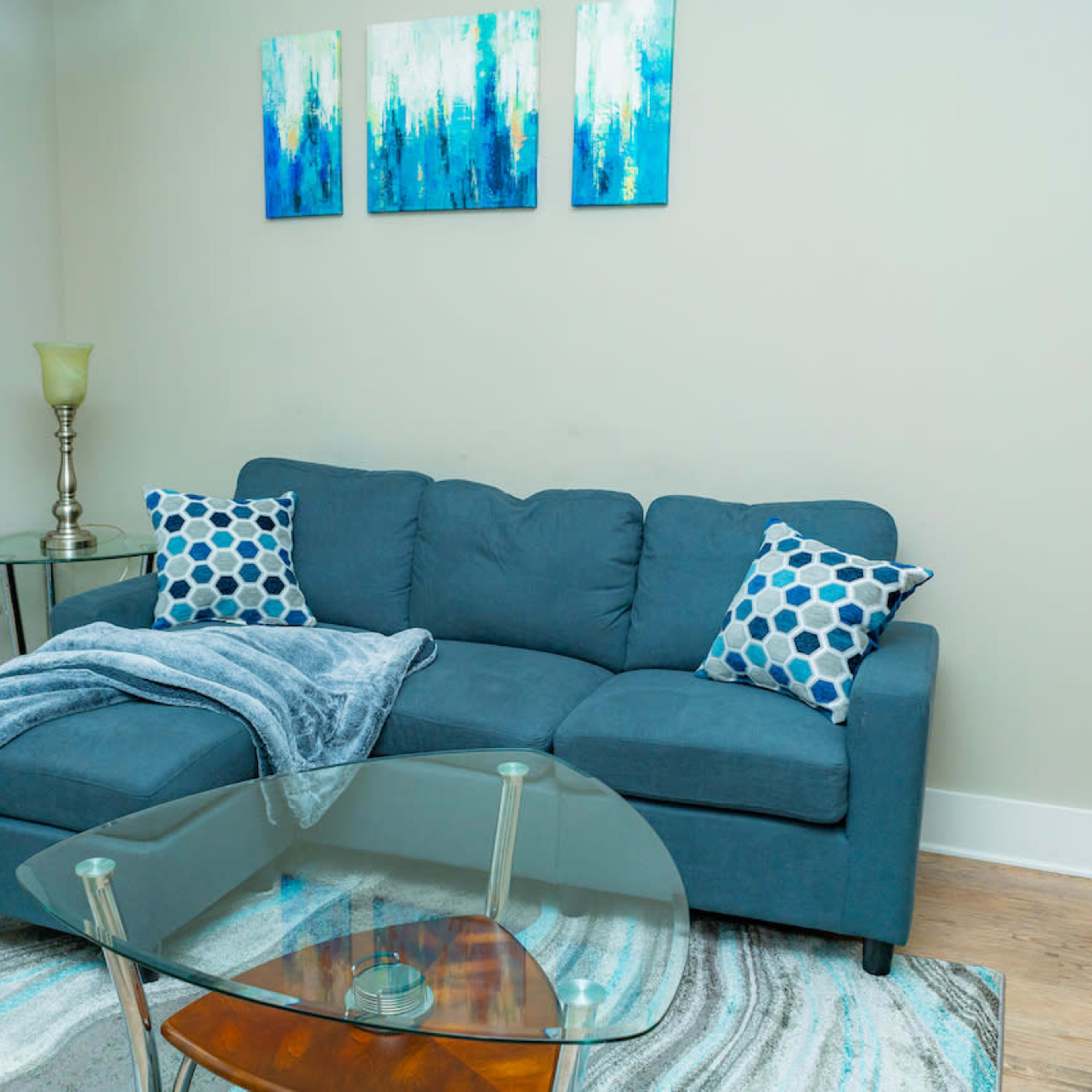 A blue couch with patterned pillows is positioned next to a glass coffee table on a patterned rug beneath three decorative blue and green paintings.
