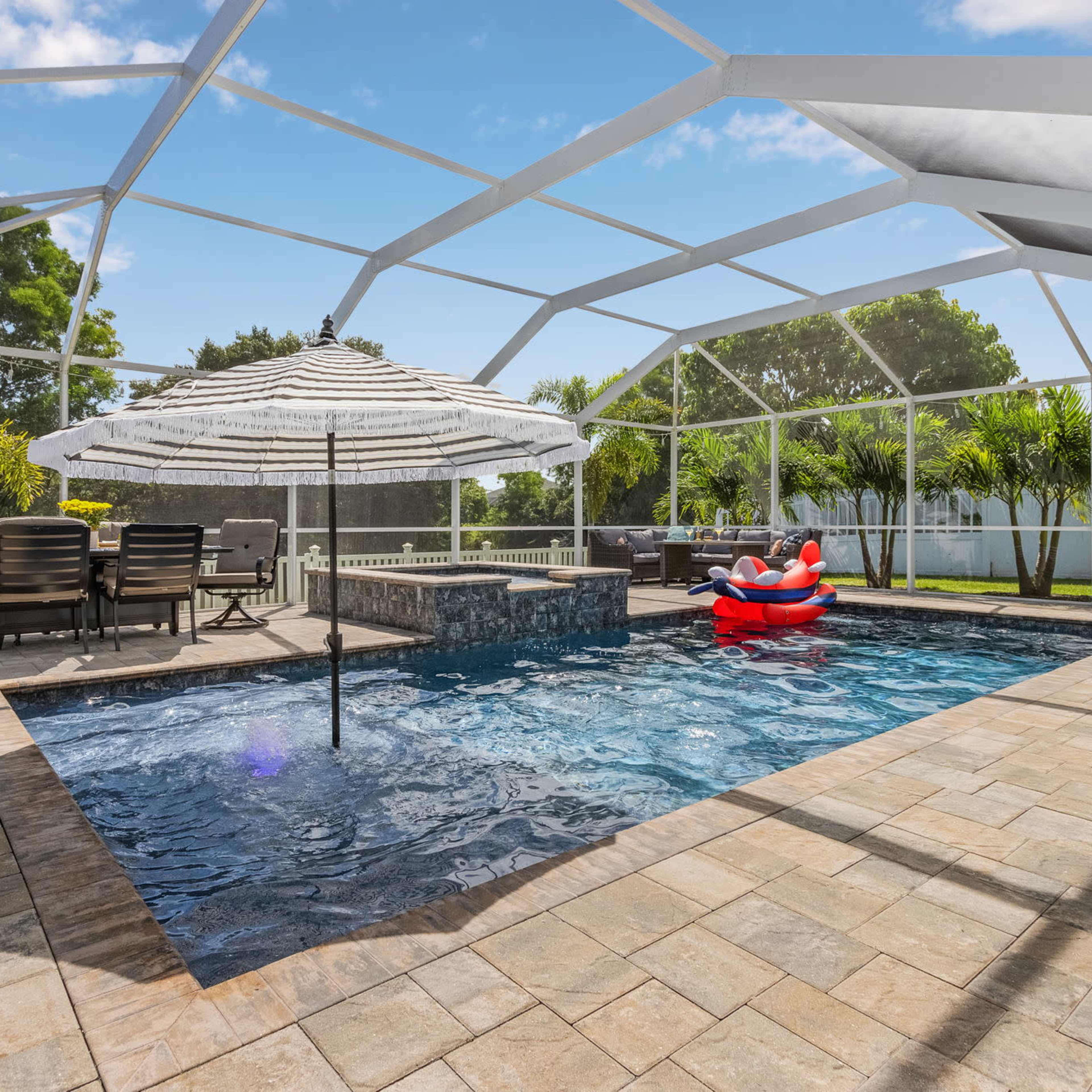 An outdoor pool area with a shaded umbrella, seating table, and a floating inflatable in a screened enclosure.