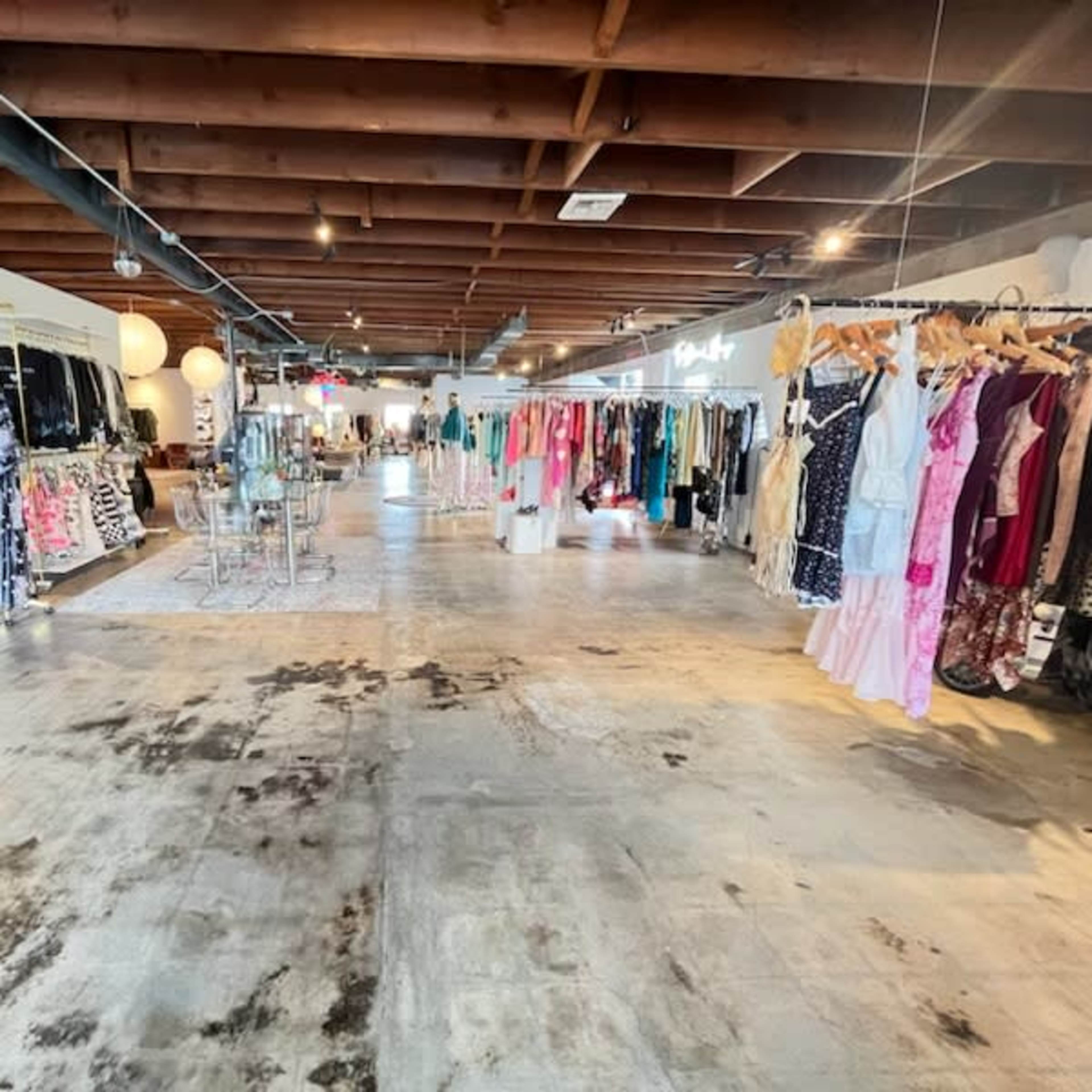 An interior retail space featuring rows of hanging clothing displays on either side, with a spacious, concrete floor and overhead wooden beams.