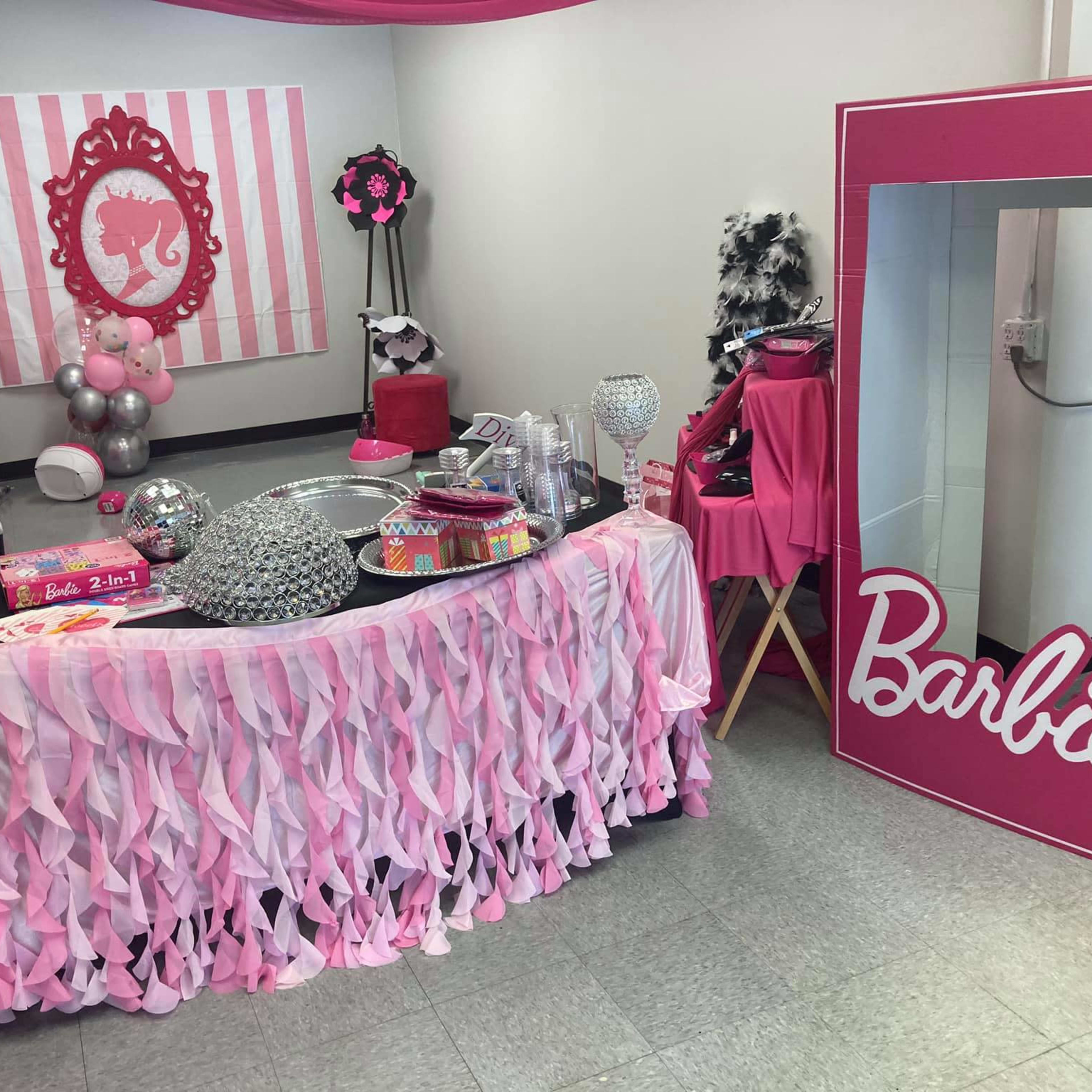 A decorated party space featuring a table draped in pink fabric, adorned with various party supplies and a large mirror with the name "Barbie" displayed prominently.