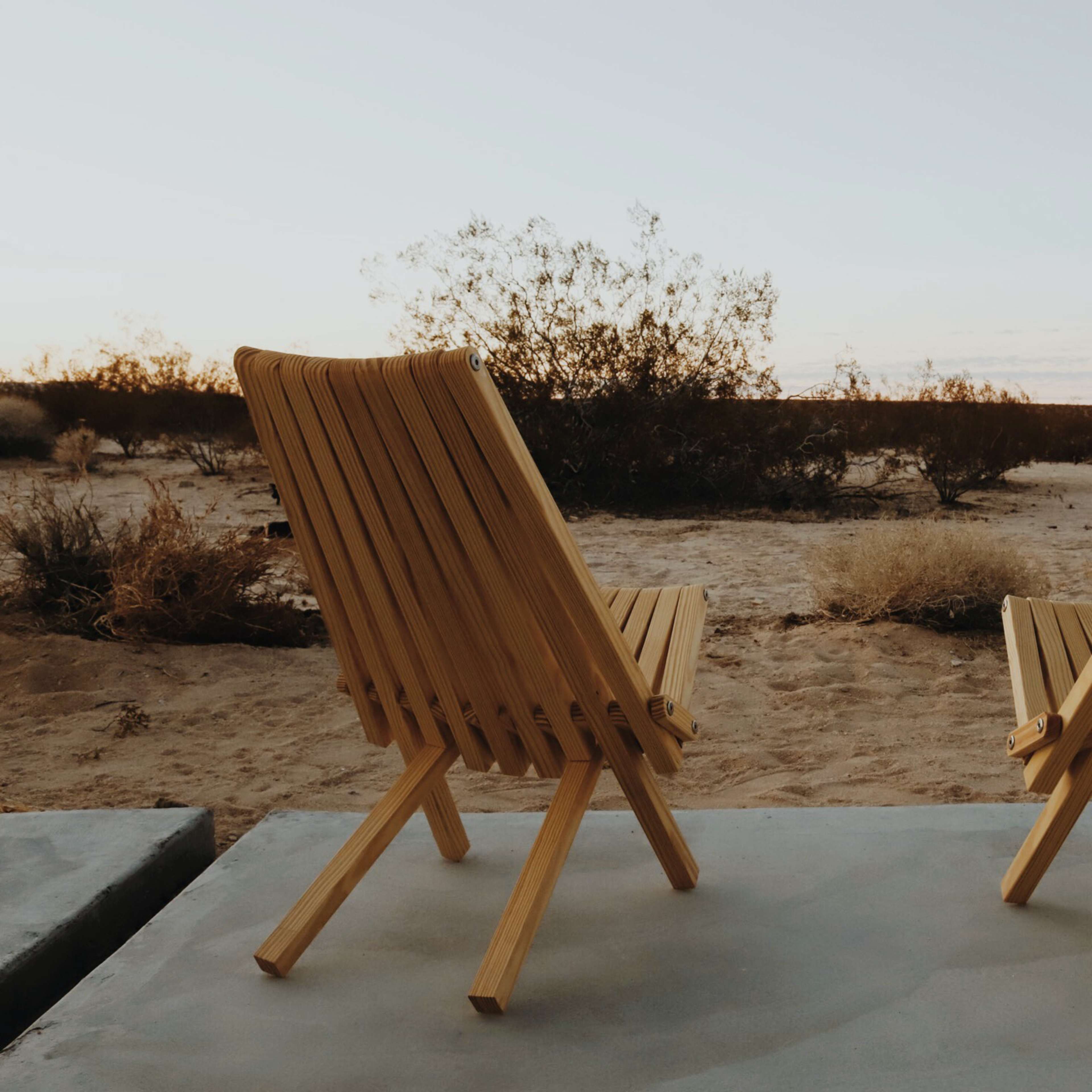 Two wooden chairs are positioned on a concrete patio, facing a sparse desert landscape under a clear sky.