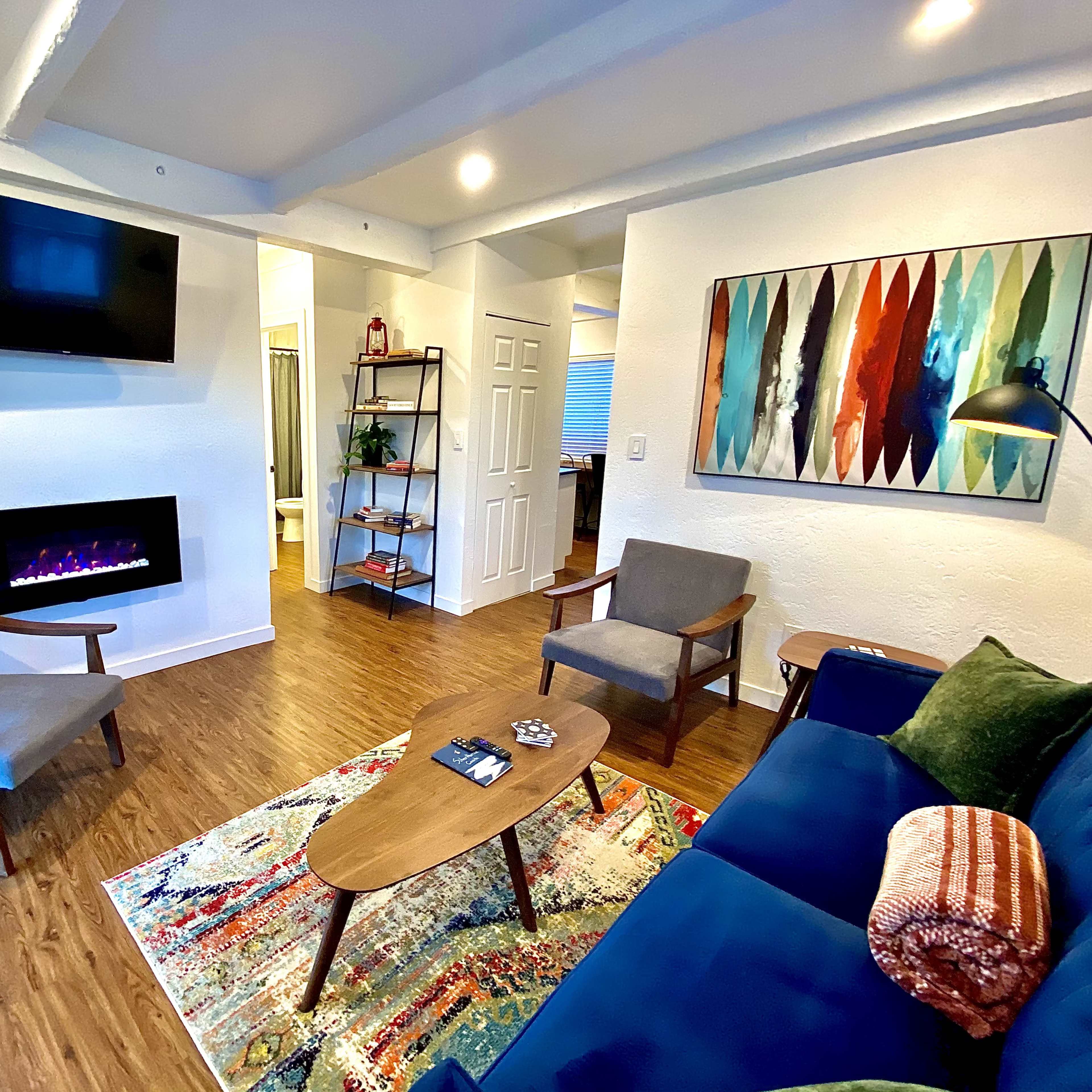 The living room features a blue couch, two gray chairs, a wooden coffee table on a patterned rug, a wall-mounted TV, and an electric fireplace.