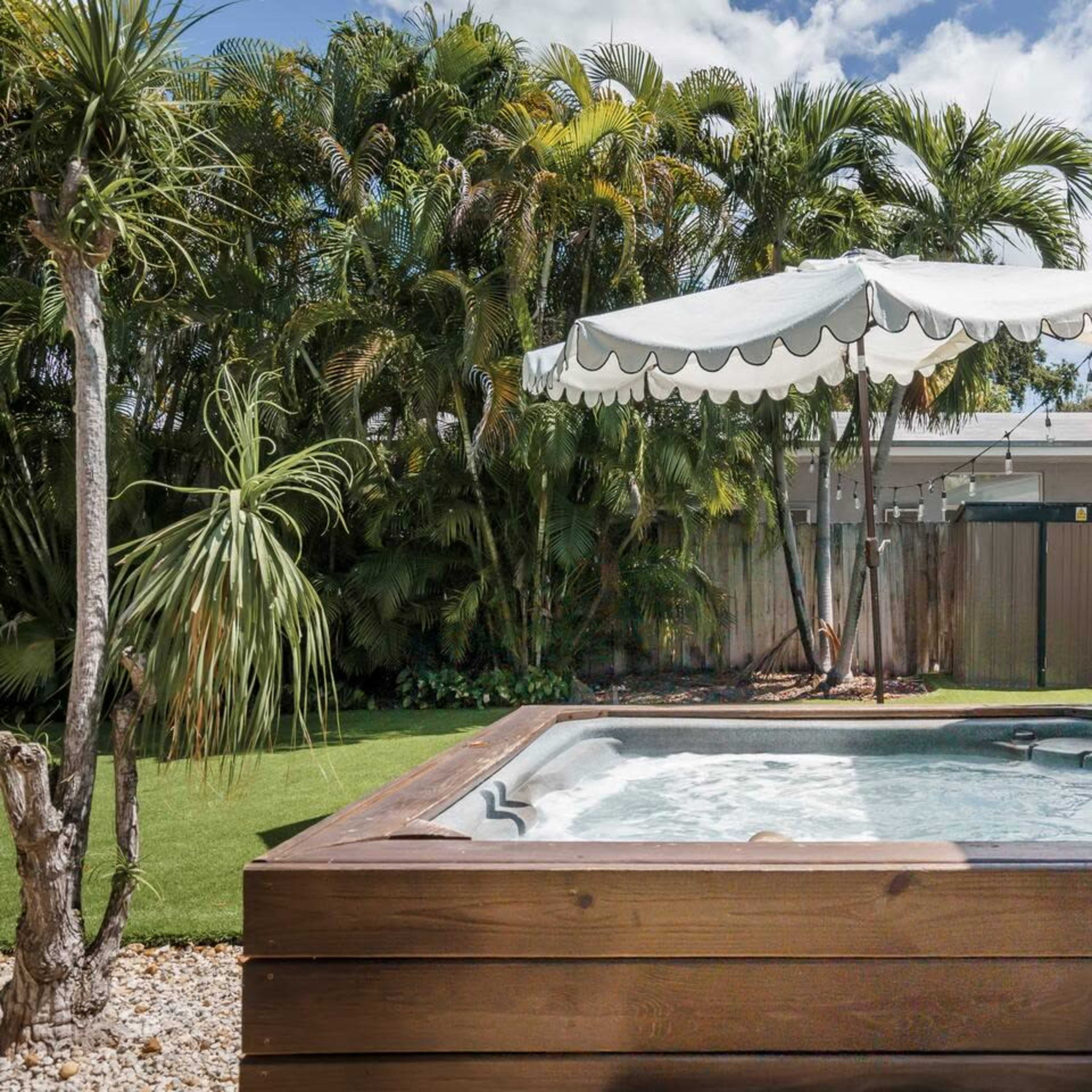 A wooden hot tub situated next to a green lawn and a white parasol surrounded by lush palm trees.