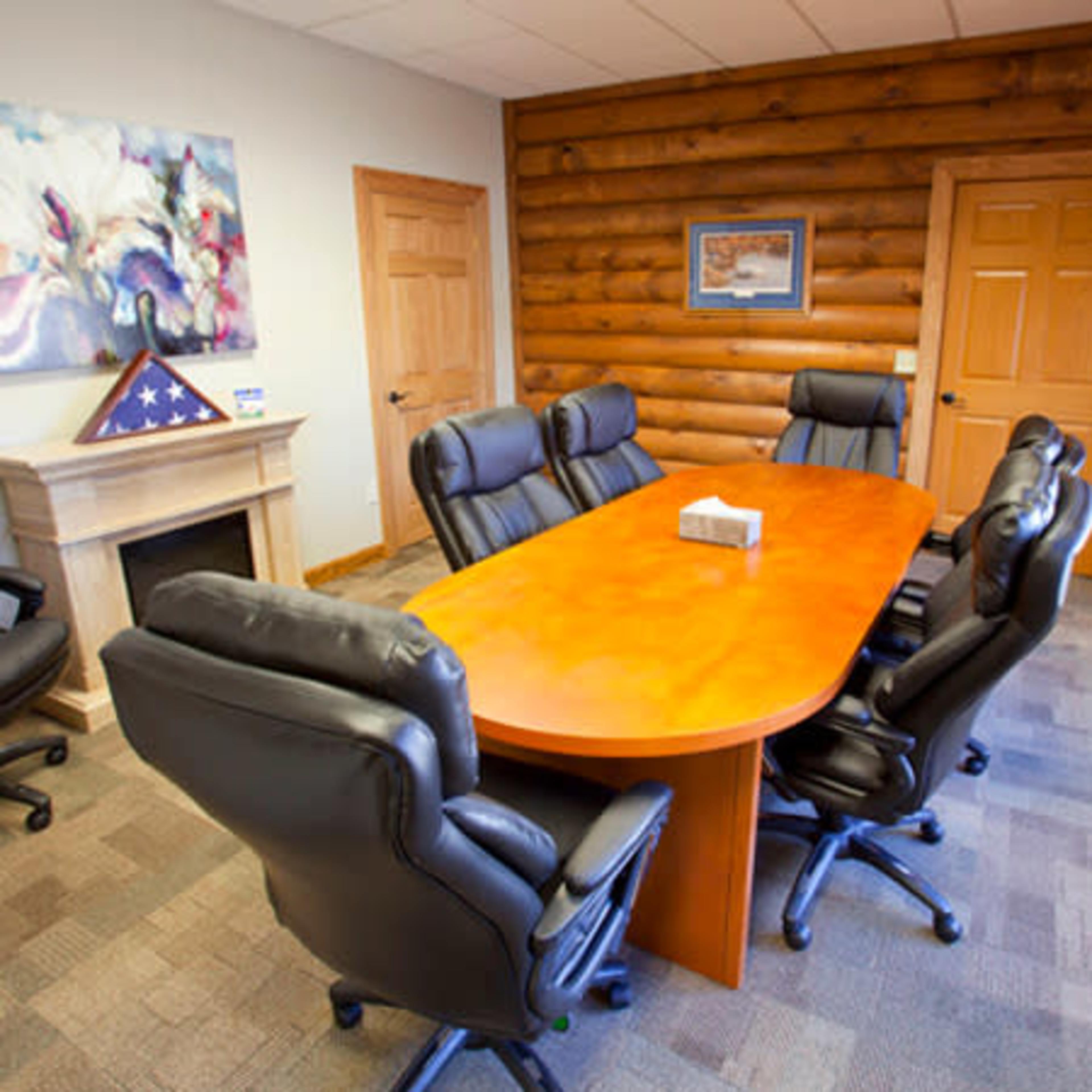 A conference room with a large wooden oval table surrounded by eight black leather office chairs, set against a log cabin-style wall.
