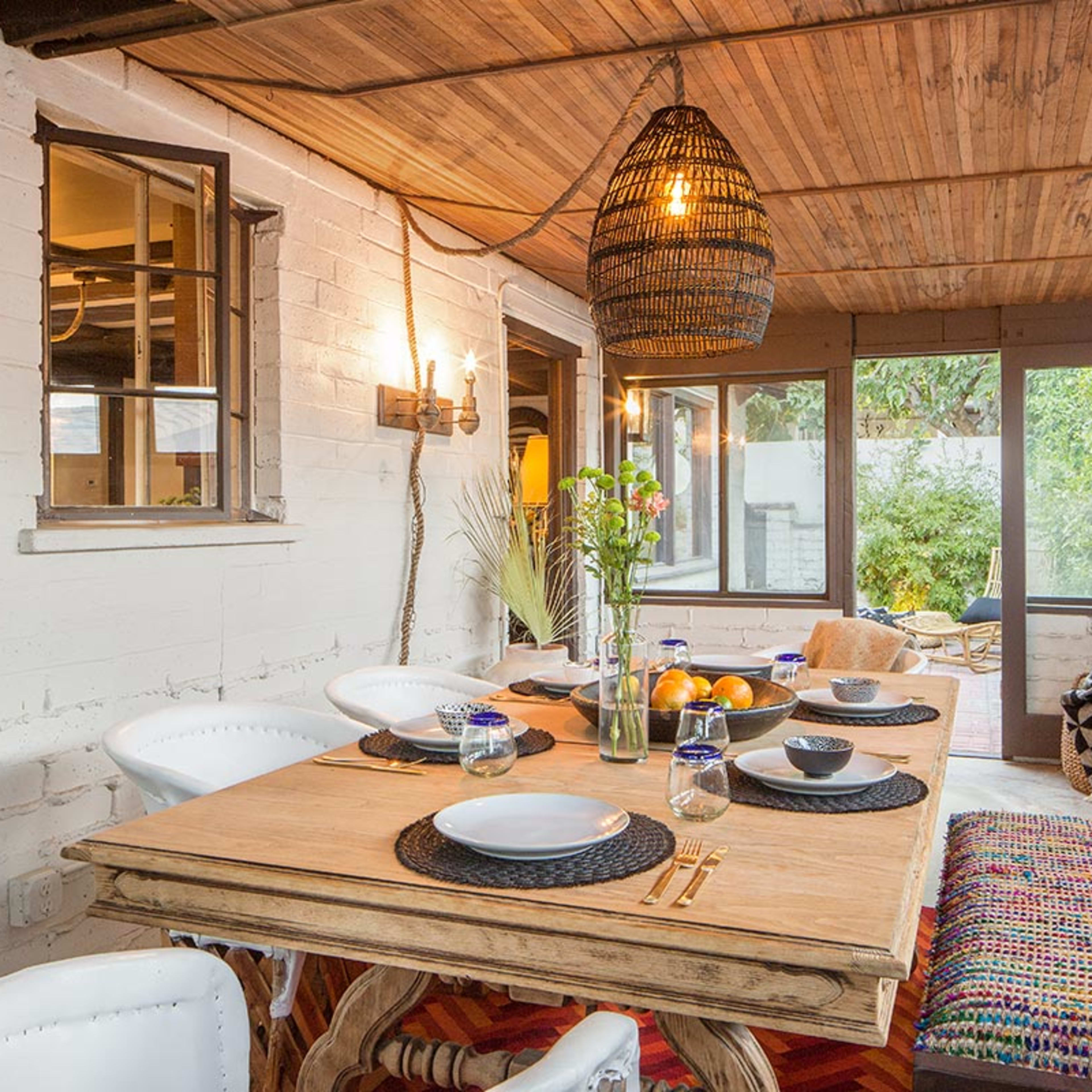 A sunlit indoor dining area with a wooden table set for a meal, surrounded by white chairs and decorated with plants and a woven light fixture.