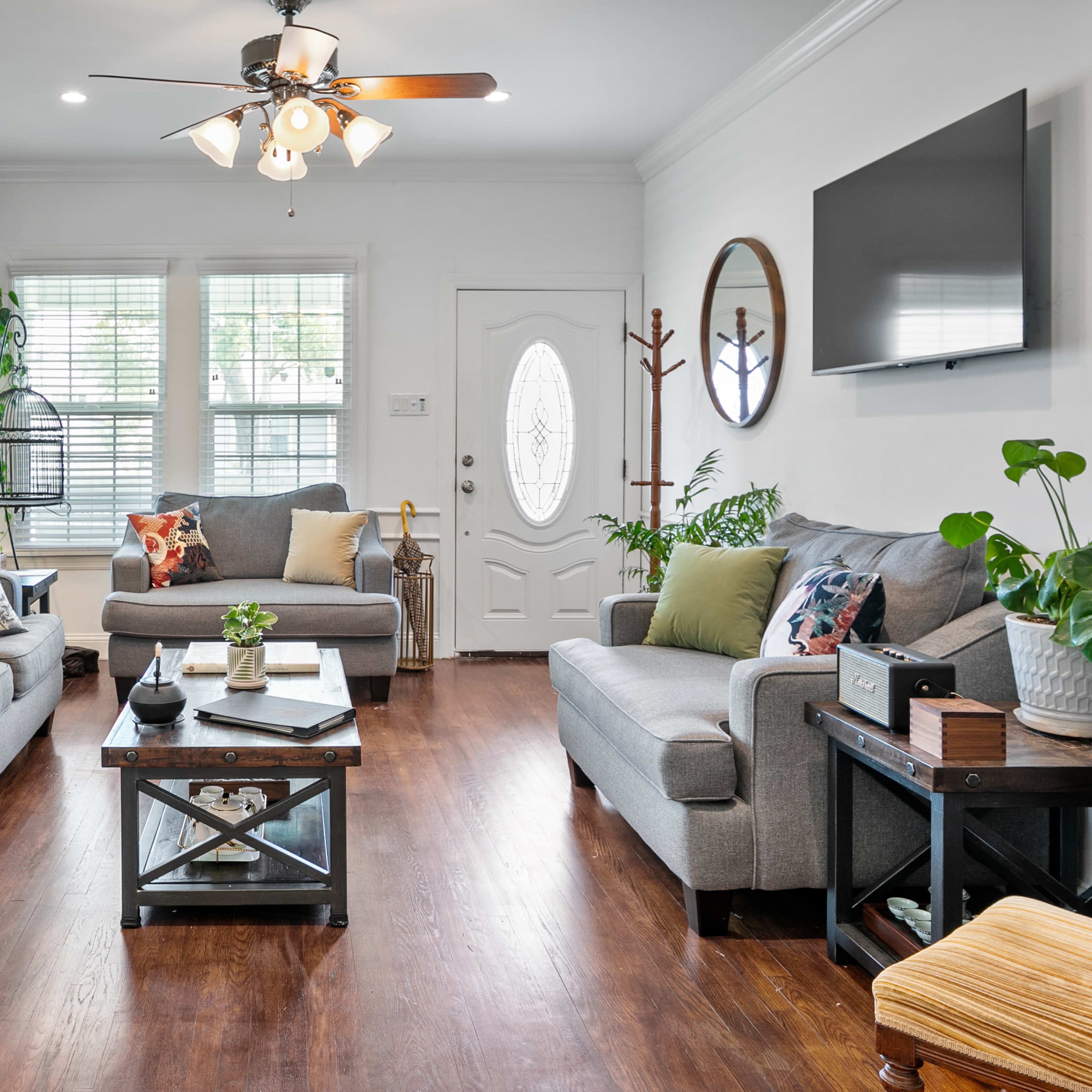 The living room features two gray sofas, a coffee table, potted plants, and a large window with natural light.